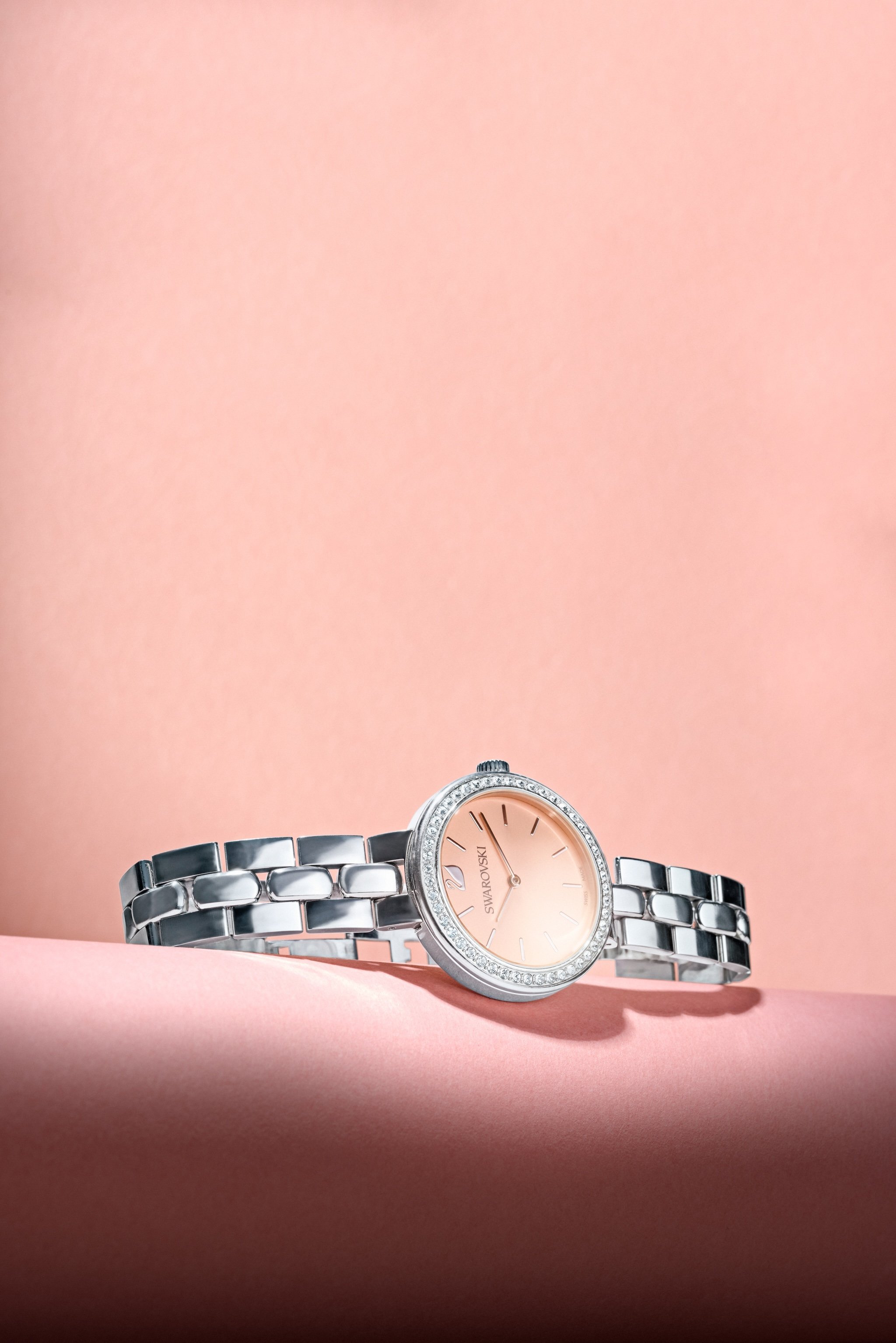 A silver Swarovski wristwatch with a pink face, placed on a pink surface against a pink background.