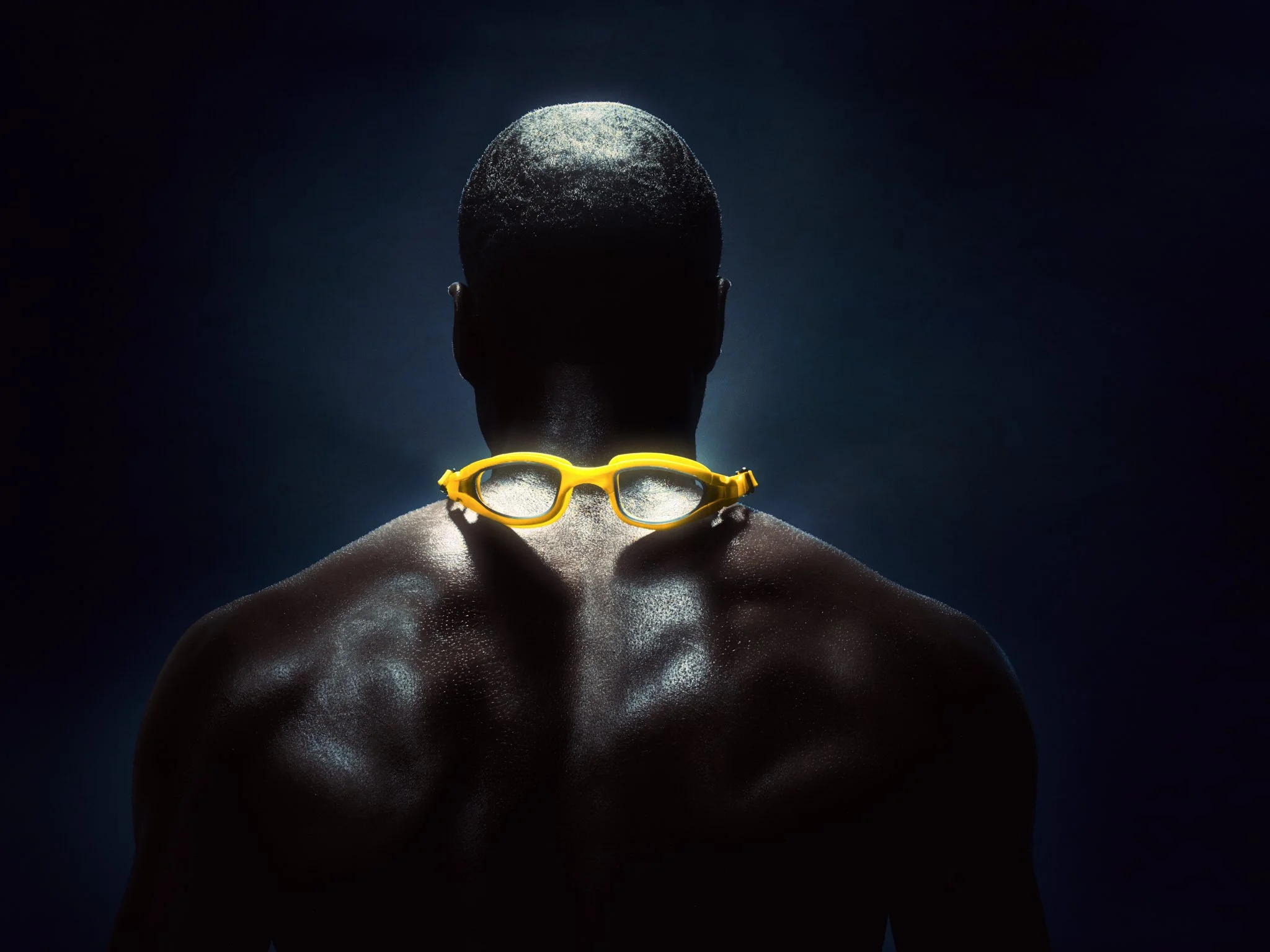 The back of a person with a shaved head and dark skin, wearing yellow swimming goggles around their neck, standing in a dark environment with water droplets on their skin.