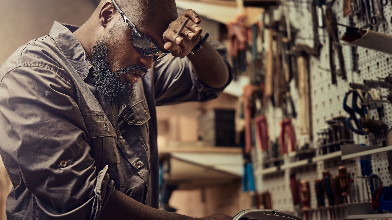A man in safety glasses working in a workshop, wiping sweat from his forehead. Tools are organized on a pegboard in the background.