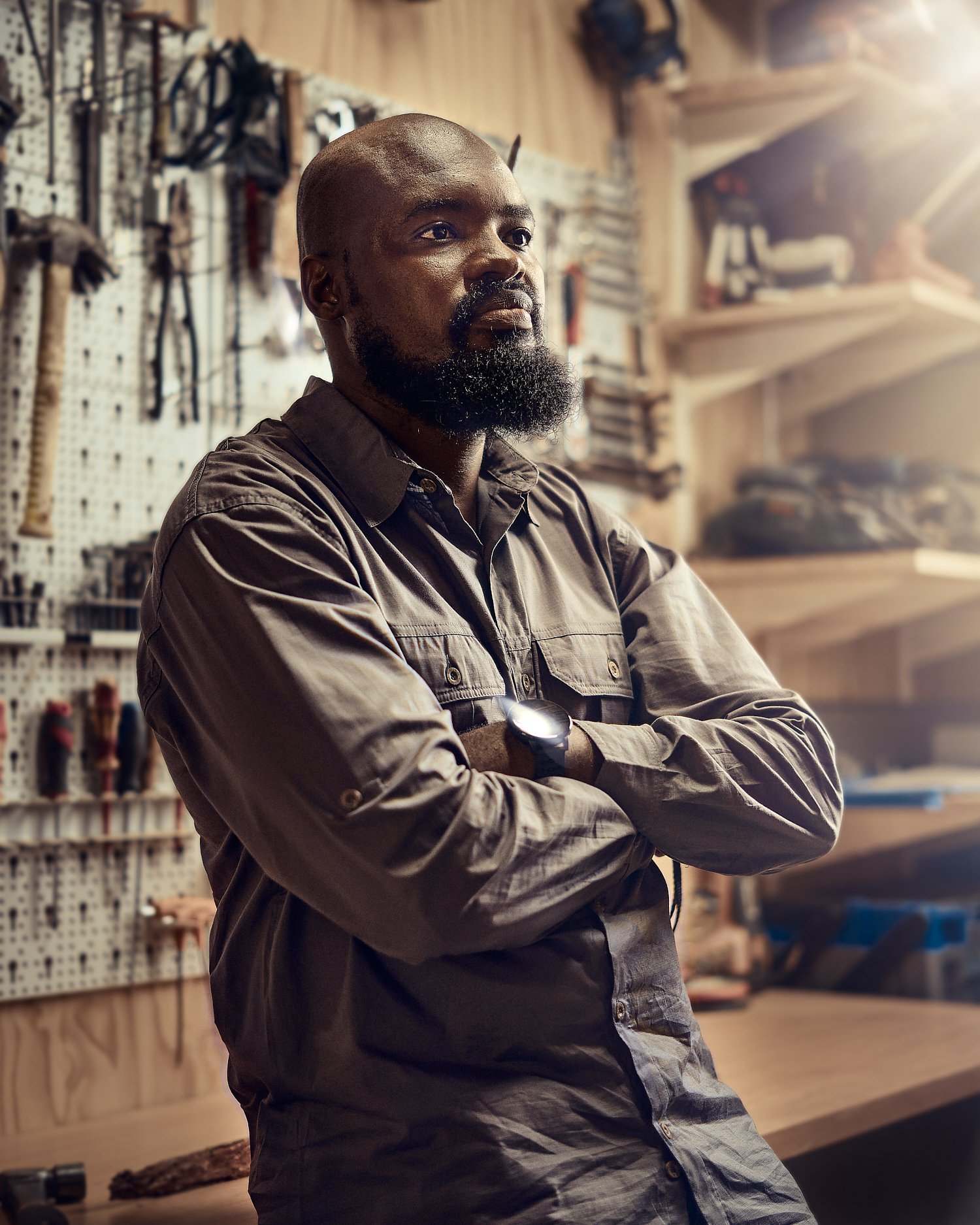 A man with a beard and work clothes standing in a workshop, arms crossed, with tools and shelves in the background.