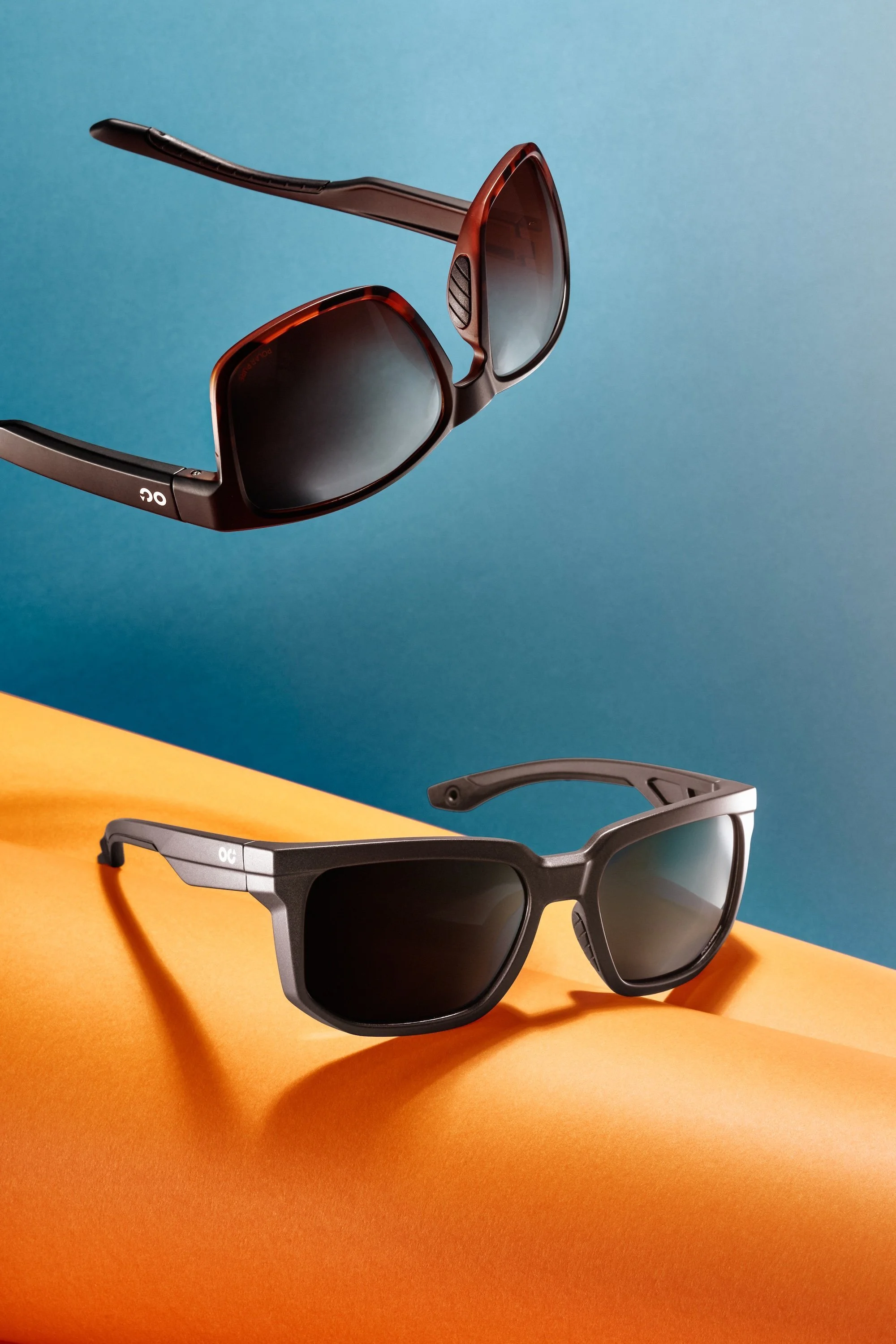 Two pairs of OC sunglasses, one black and one brown, displayed on an orange and blue background.
