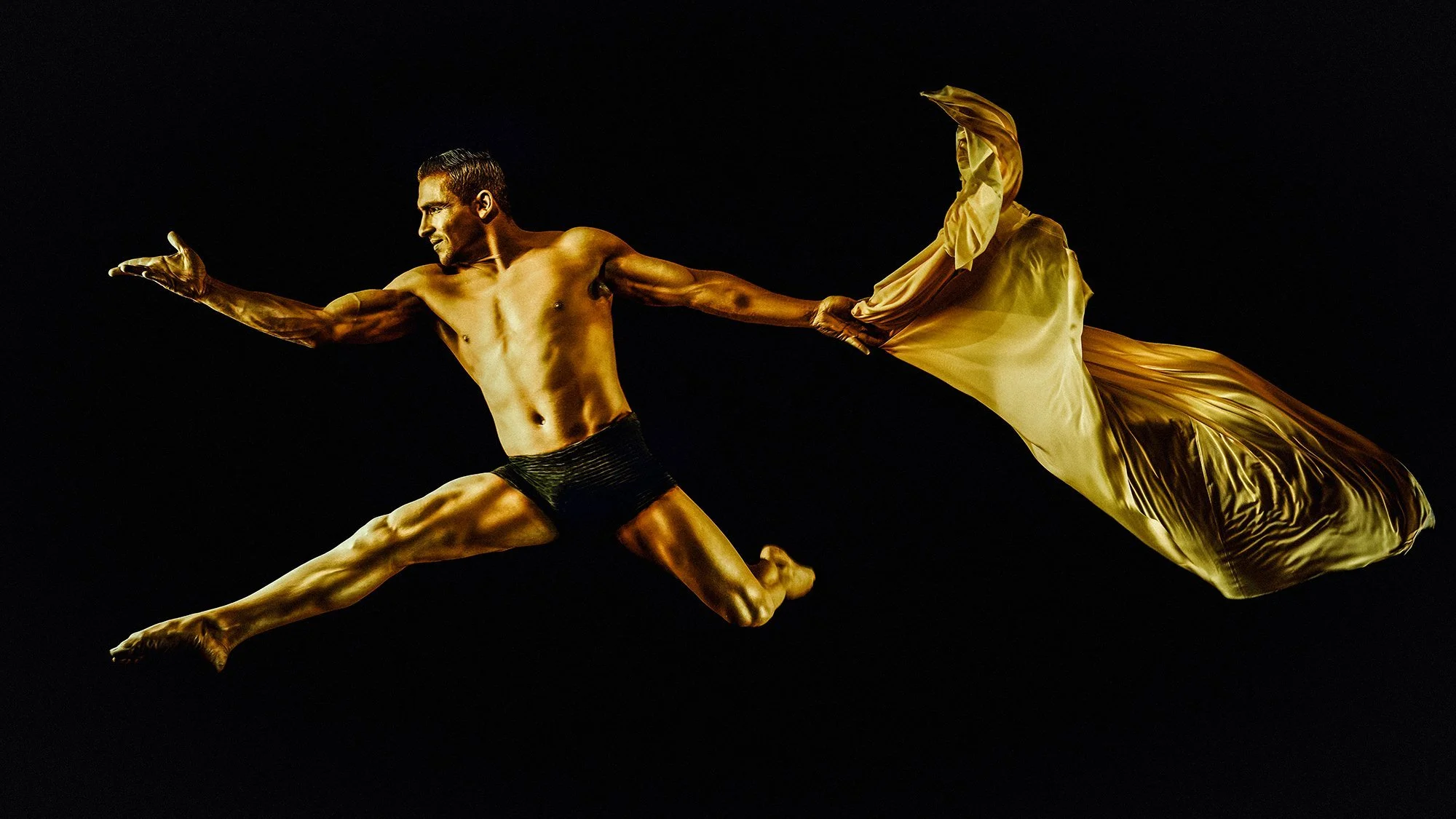Dancer in mid-air leap with golden fabric against a black background.