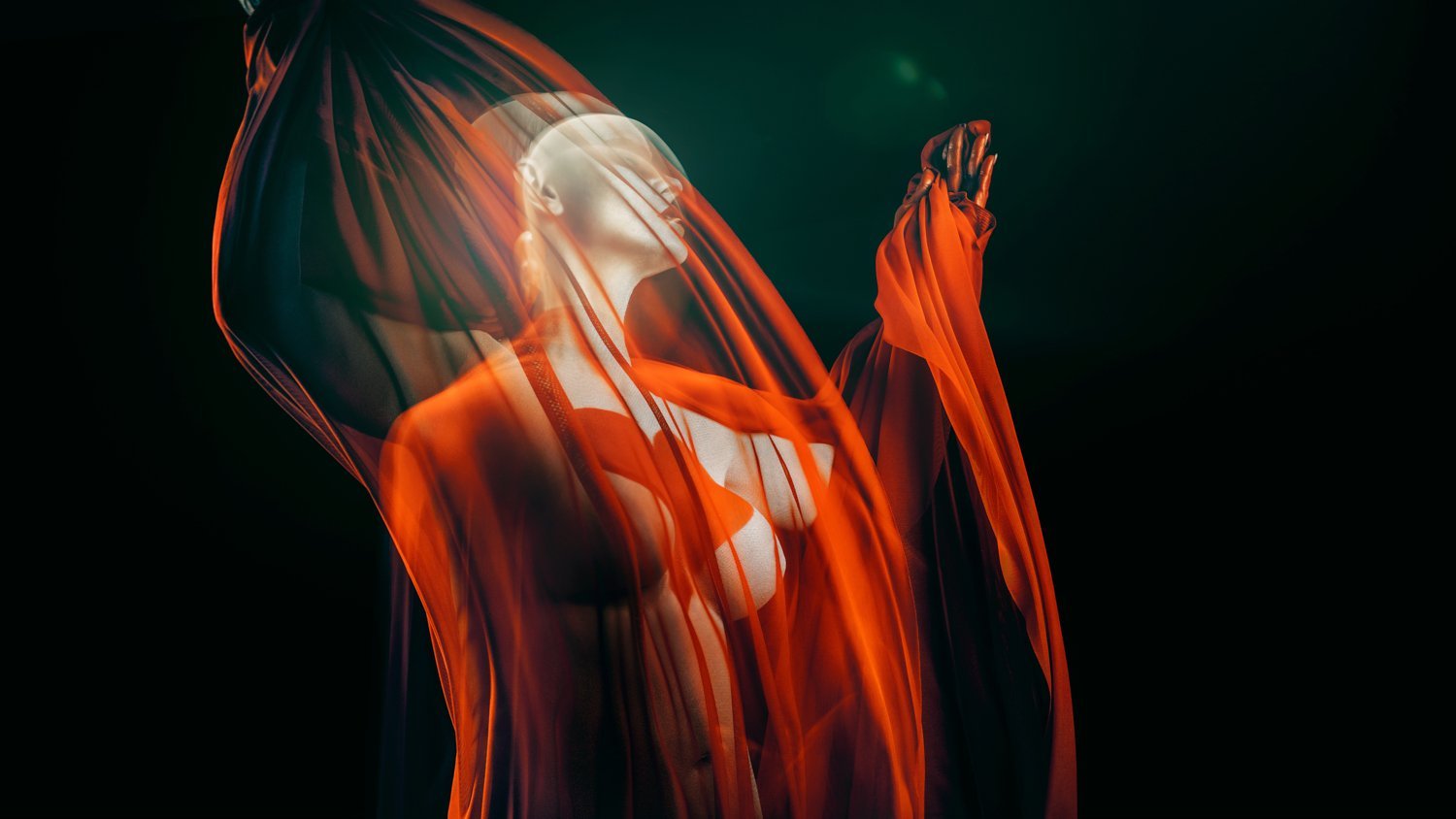 Artistic photograph of a person draped in sheer, flowing orange fabric against a dark background.