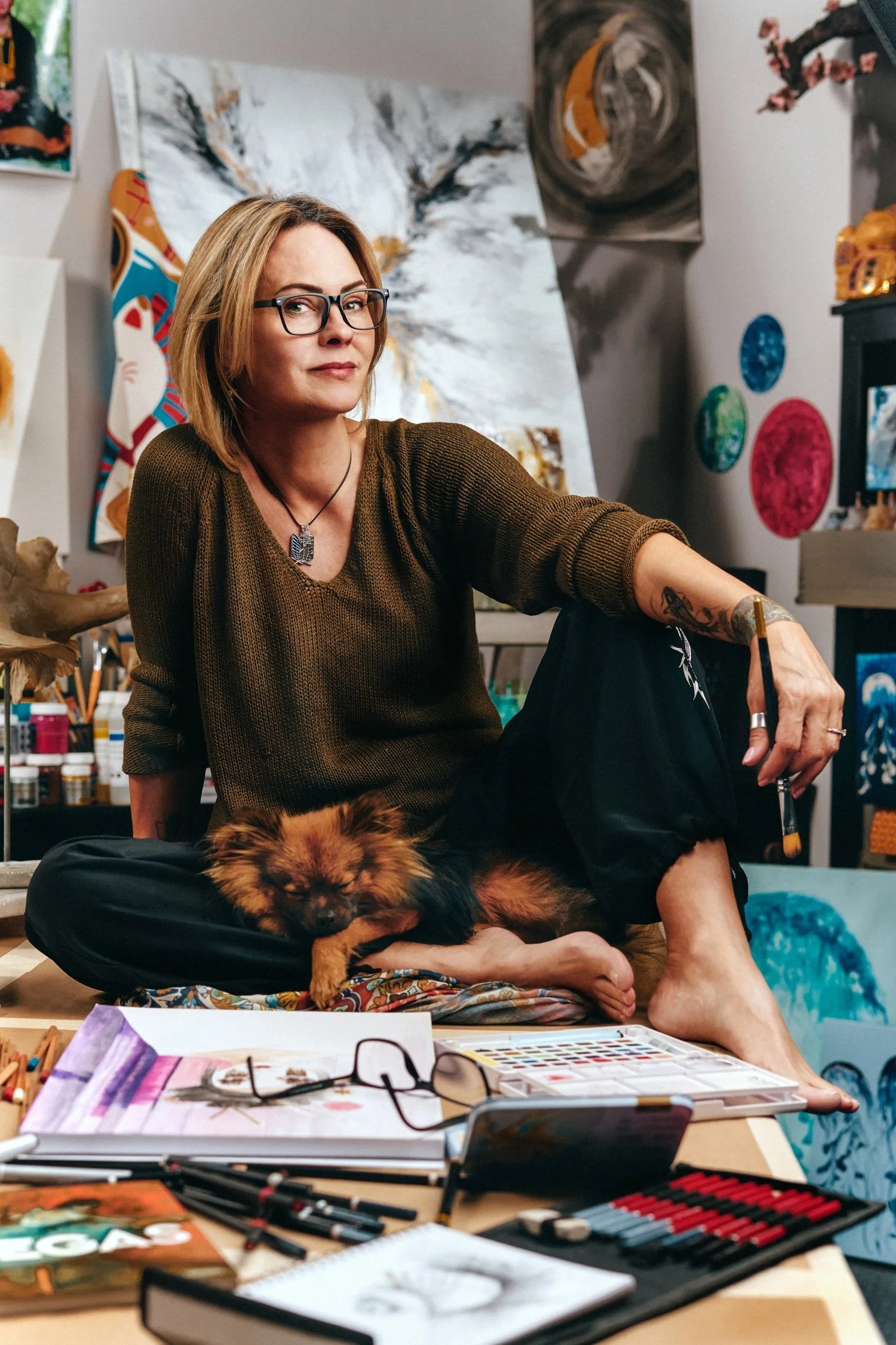 Artist sitting in a studio with a small dog on their lap, surrounded by art supplies and paintings.