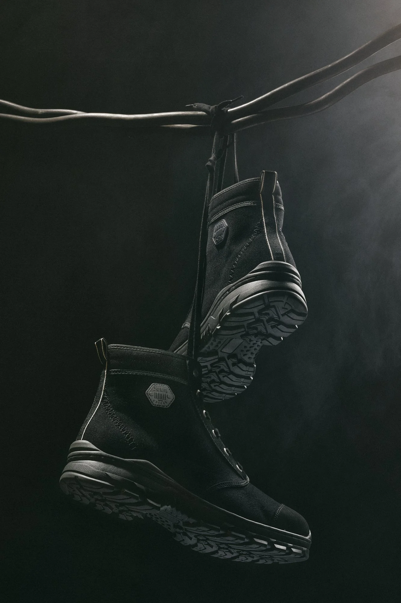 Pair of black boots with rugged soles hanging from overhead wires against a dark background.