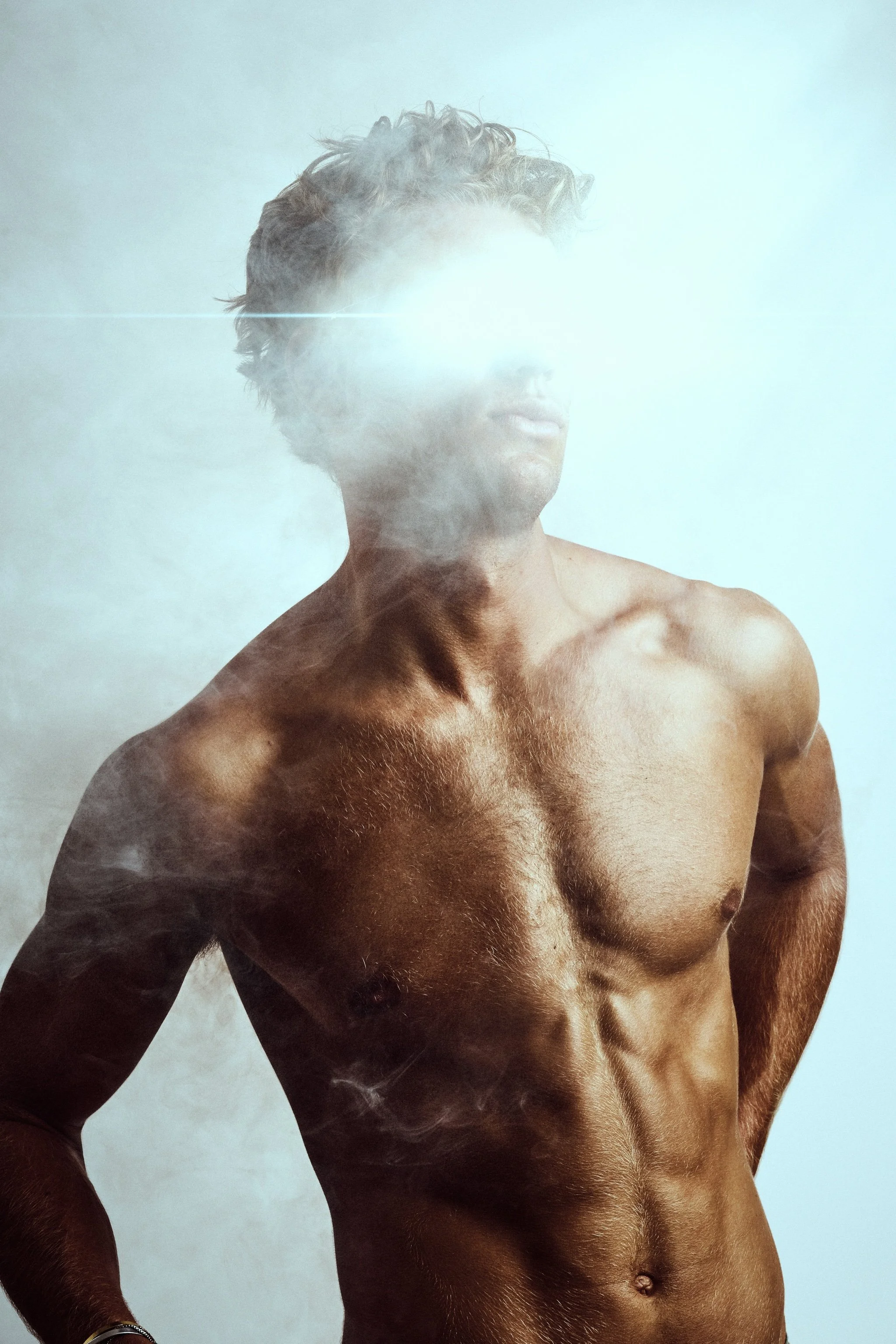 Topless muscular man with light and smoke covering his face, standing against a light background.