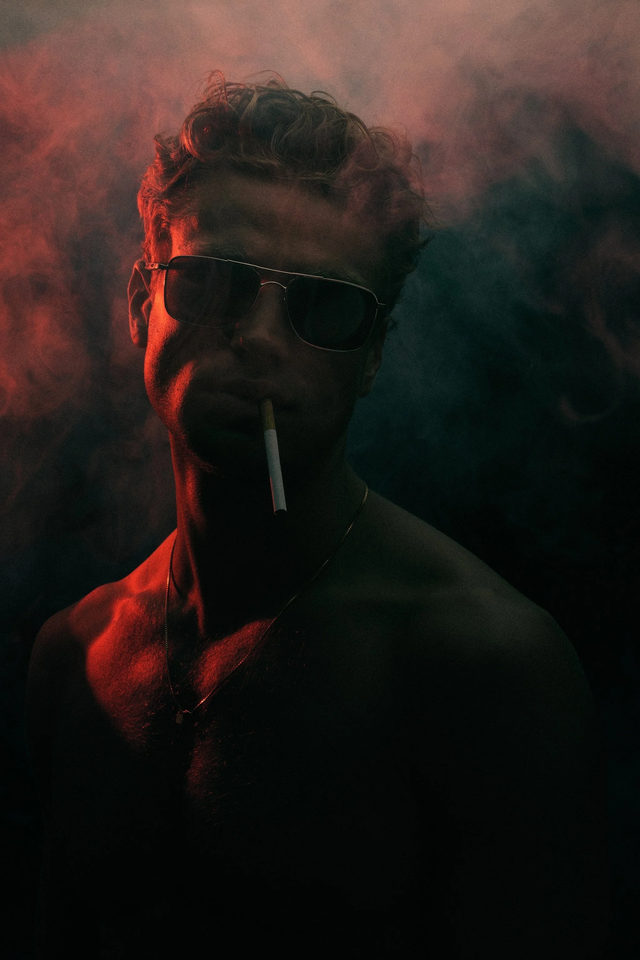 A man wearing sunglasses and a necklace, smoking a cigarette, with dramatic red and dark lighting and surrounding smoke.