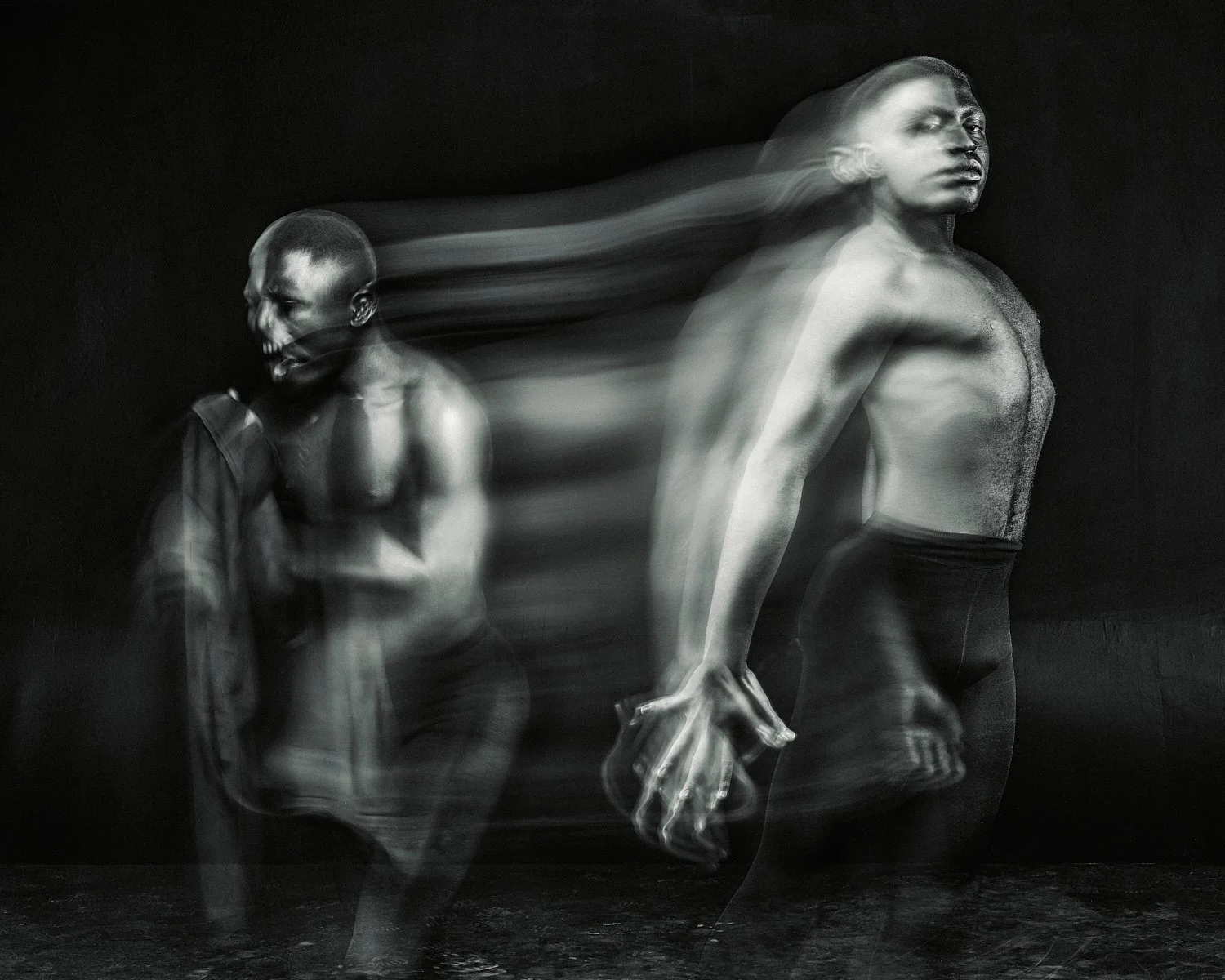 Artistic black and white photograph of two shirtless men in motion against a dark background. The image uses a long exposure effect to create a sense of fluid movement, with blurred figures and dynamic poses conveying energy and emotion.