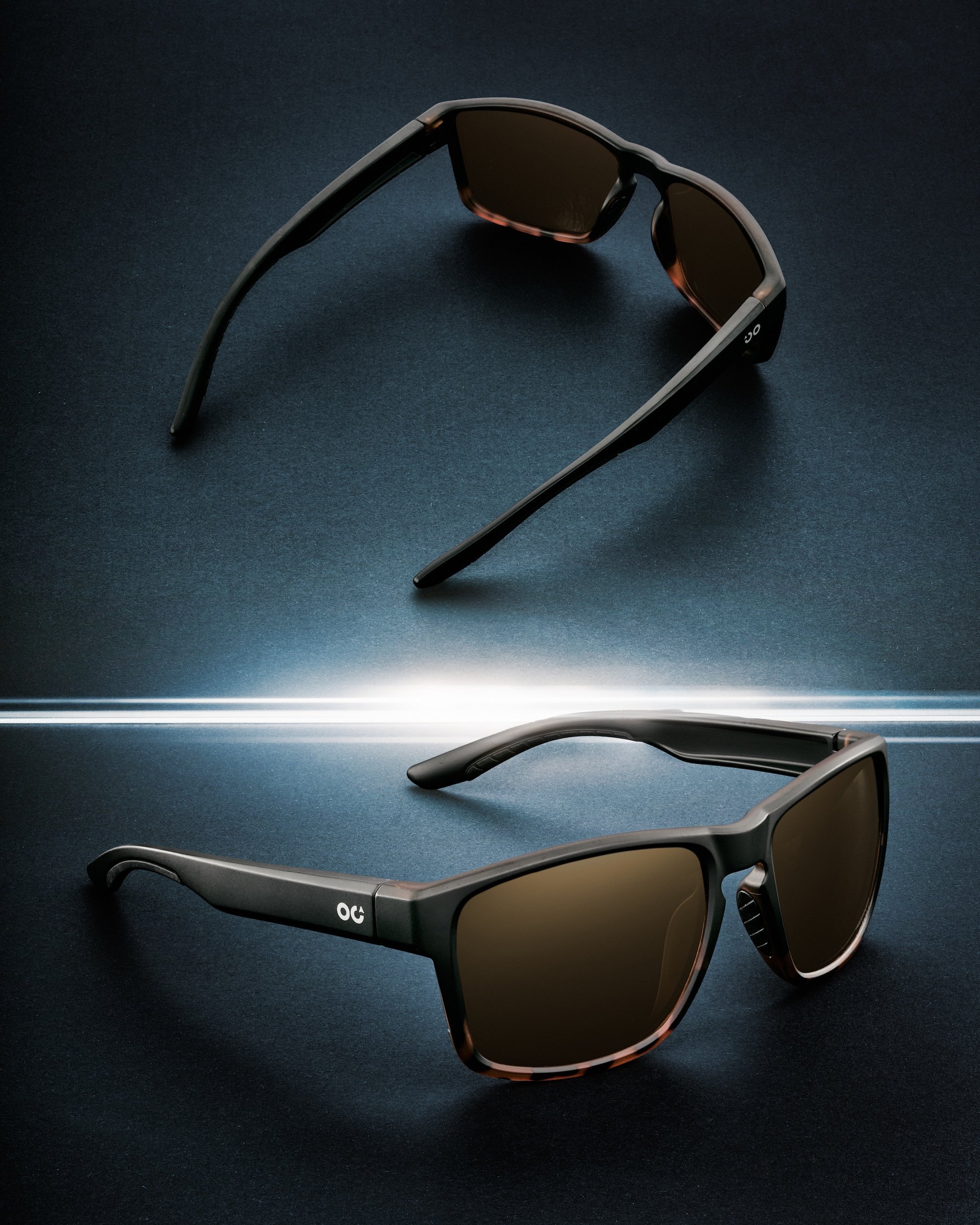 Two black sunglasses with dark lenses are placed on a dark surface, illuminated by a bright horizontal light in the middle, featuring a sleek and modern design.