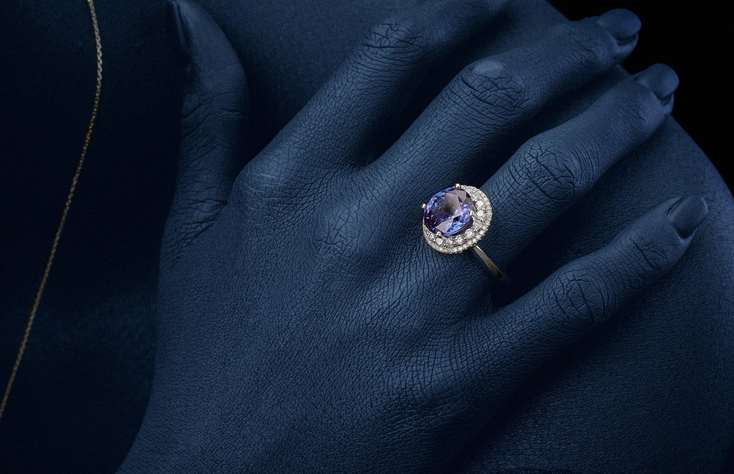 Close-up of a hand with blue skin, wearing a silver ring with a large tanzanite and surrounded by small clear diamonds, resting on a model's shoulder.