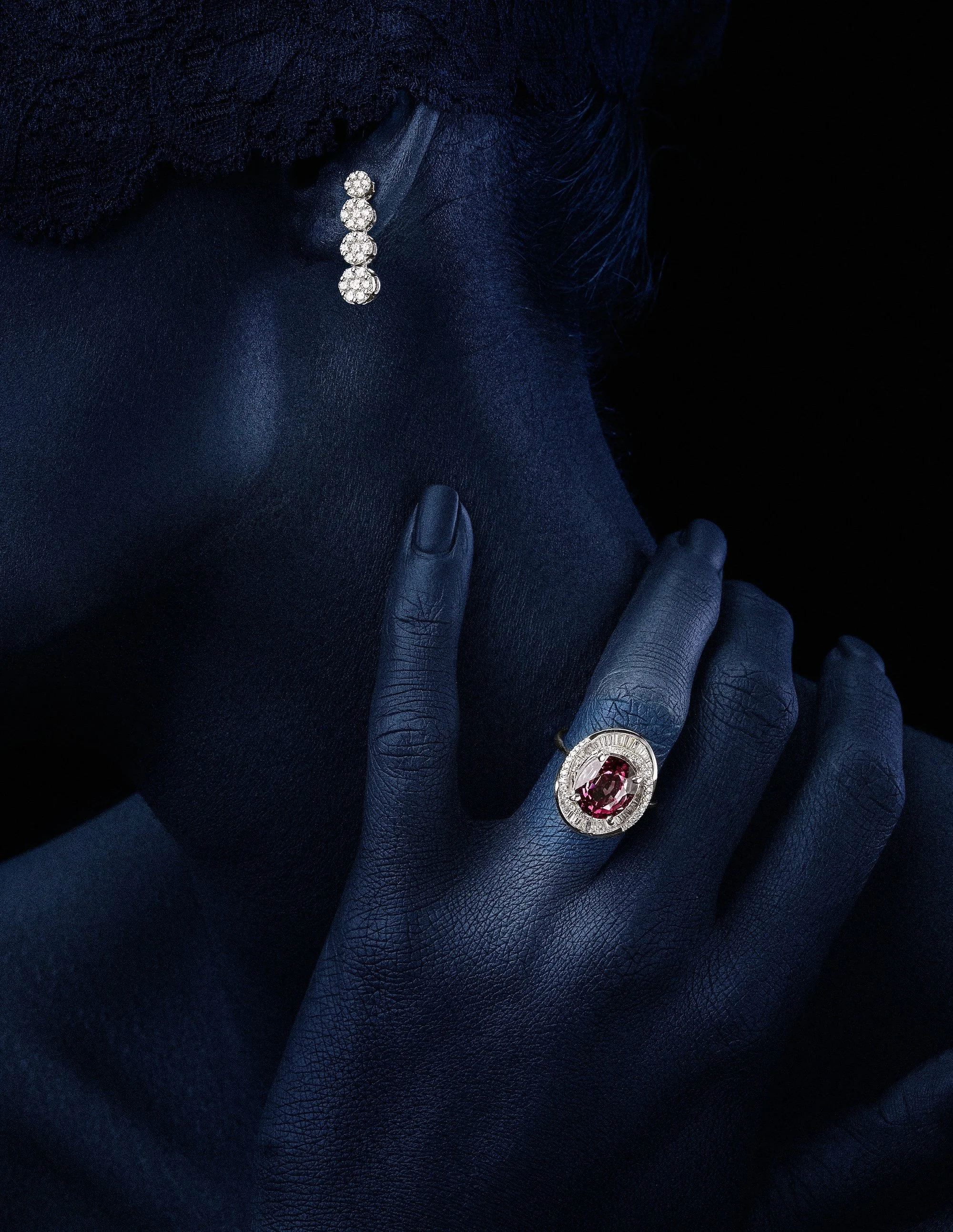 Close-up of a hand with dark blue skin, wearing a large ring with a red gemstone and surrounded by smaller clear stones, resting on the face of a person with dark blue skin, wearing a dark fabric covering part of their face, all against a black backg