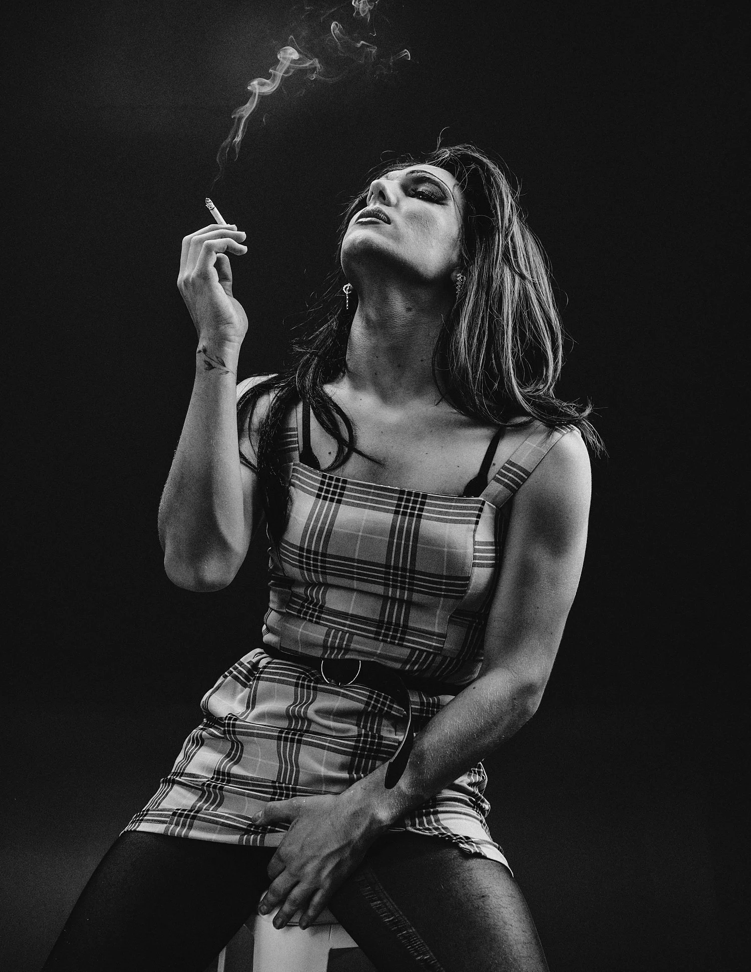 Black and white photo of a person with long hair wearing a plaid dress, sitting and smoking a cigarette.