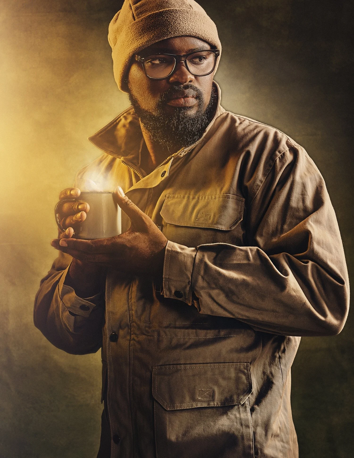 A person wearing glasses, a beanie, and a brown jacket, holding a steaming mug against a warm-toned background.