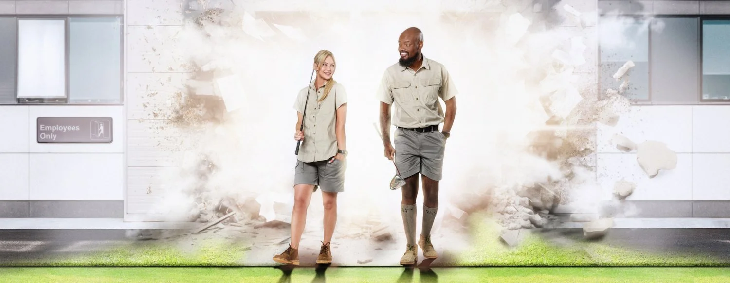 A man and woman in safari outfits joyfully walking on grass with golf clubs; background shows an "Employees Only" sign, a door bursting open with debris.