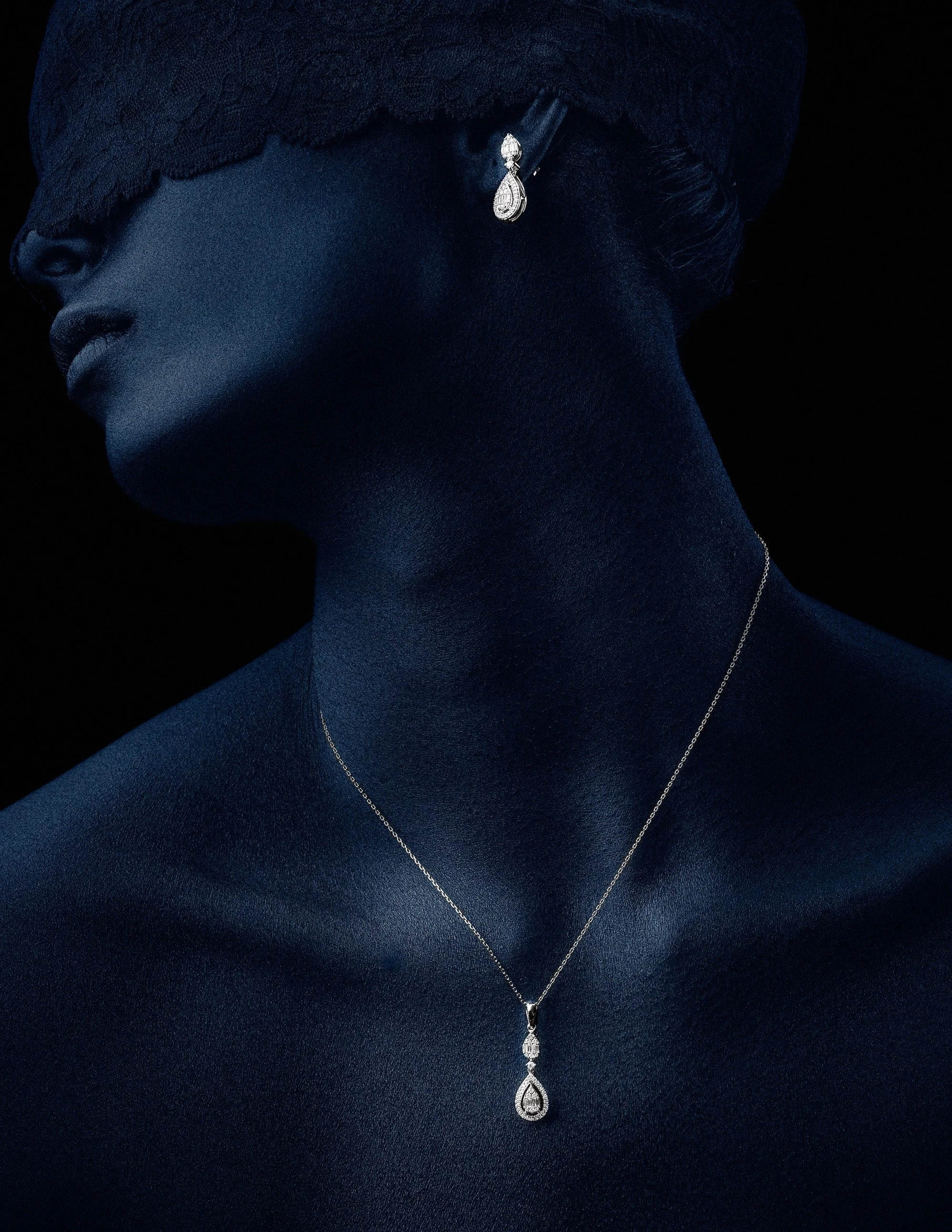 A woman with dark skin wearing diamond jewelry, including a pair of dangling earrings and a necklace with a teardrop-shaped pendant, against a black background.