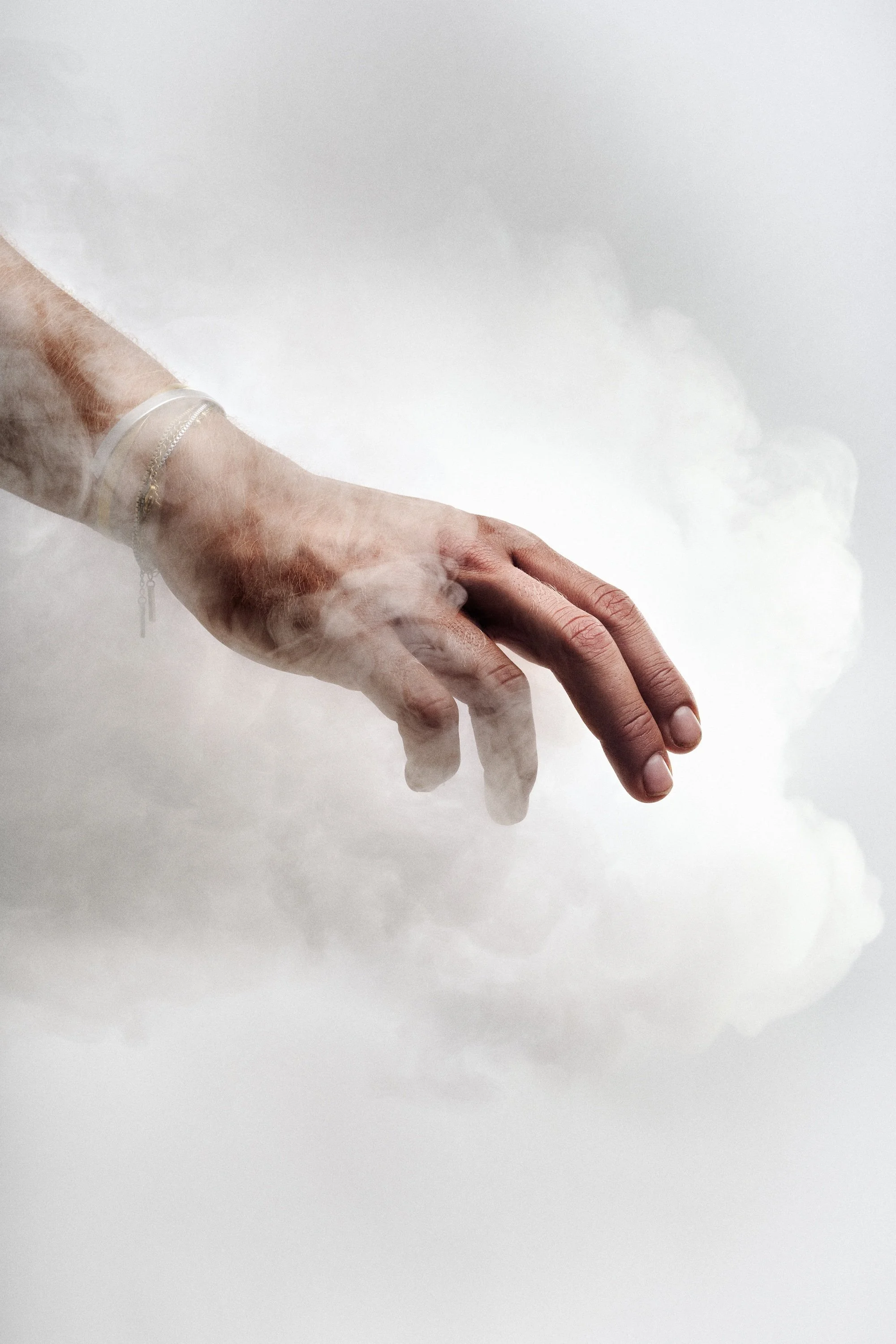 A human hand reaching out through a cloud of smoke or fog, wearing a bracelet on the wrist.