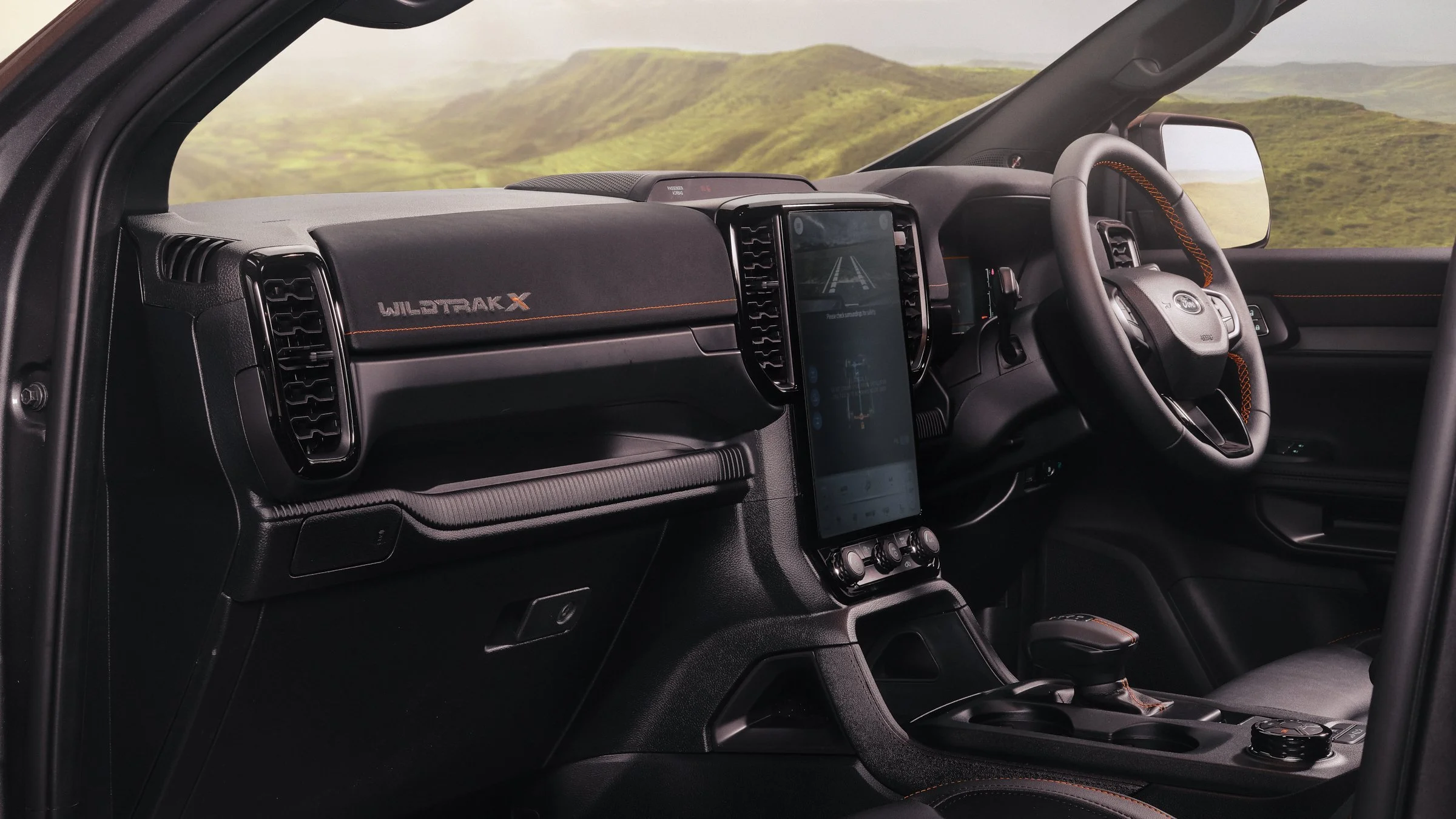 Interior of a Ford vehicle with a digital dashboard, steering wheel, and large touchscreen display in the center, against a backdrop of green hills visible through the windshield.