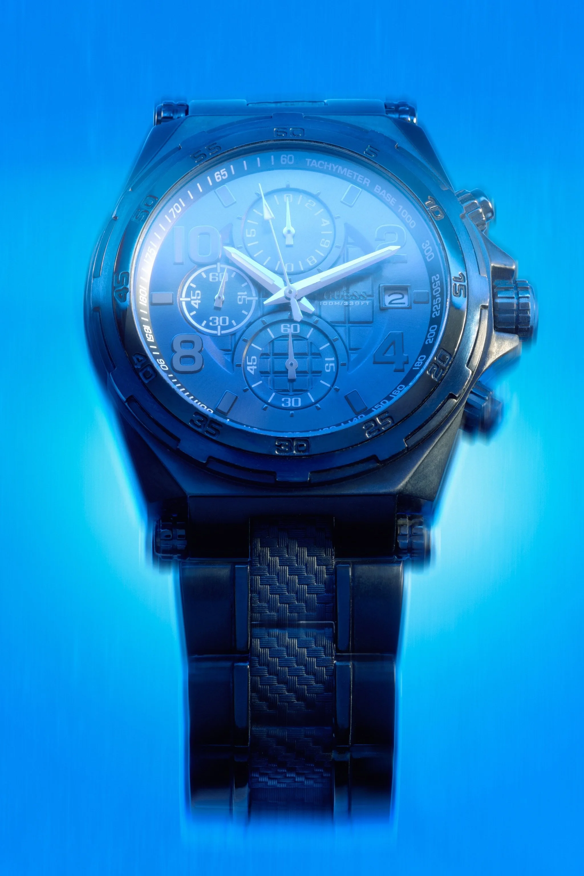 A close-up of a blue men's Guess wristwatch with a tachymeter bezel, three subdials, a date window, and a metal strap, illuminated with blue lighting.