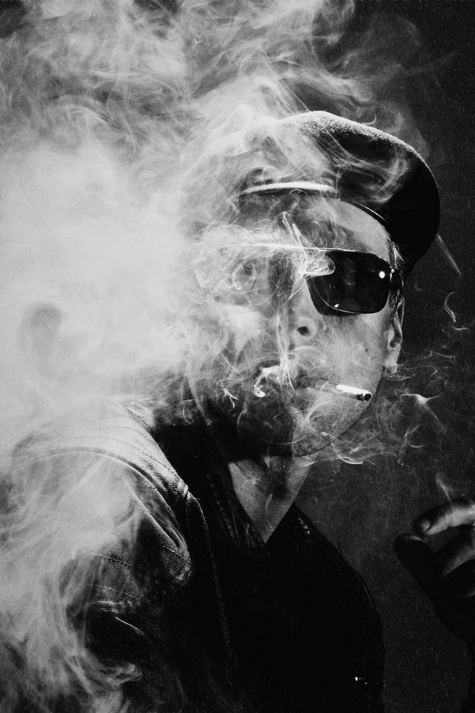 A person wearing sunglasses and a beret, smoking with smoke surrounding them, in a black and white photo. Reminiscent of a 60's James Dean rebellious portrait. 