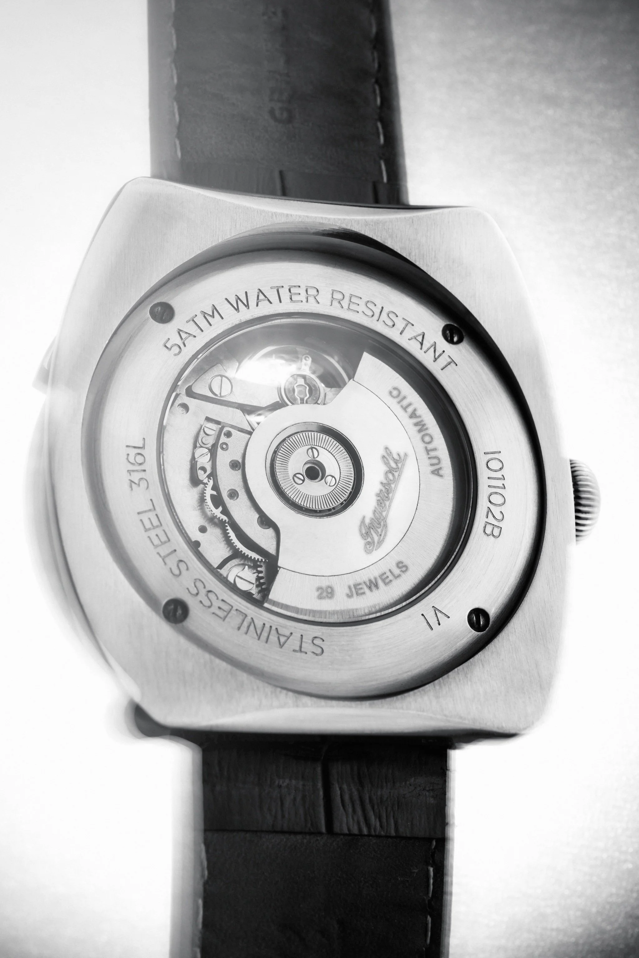 Close-up of a square-shaped wristwatch with visible mechanical movement through a transparent case back, featuring engravings such as 'STAINLESS STEEL 316L,' '5 ATM WATER RESISTANT,' '29 JEWELS,' and the brand name 'Famous.' The watch has a black str