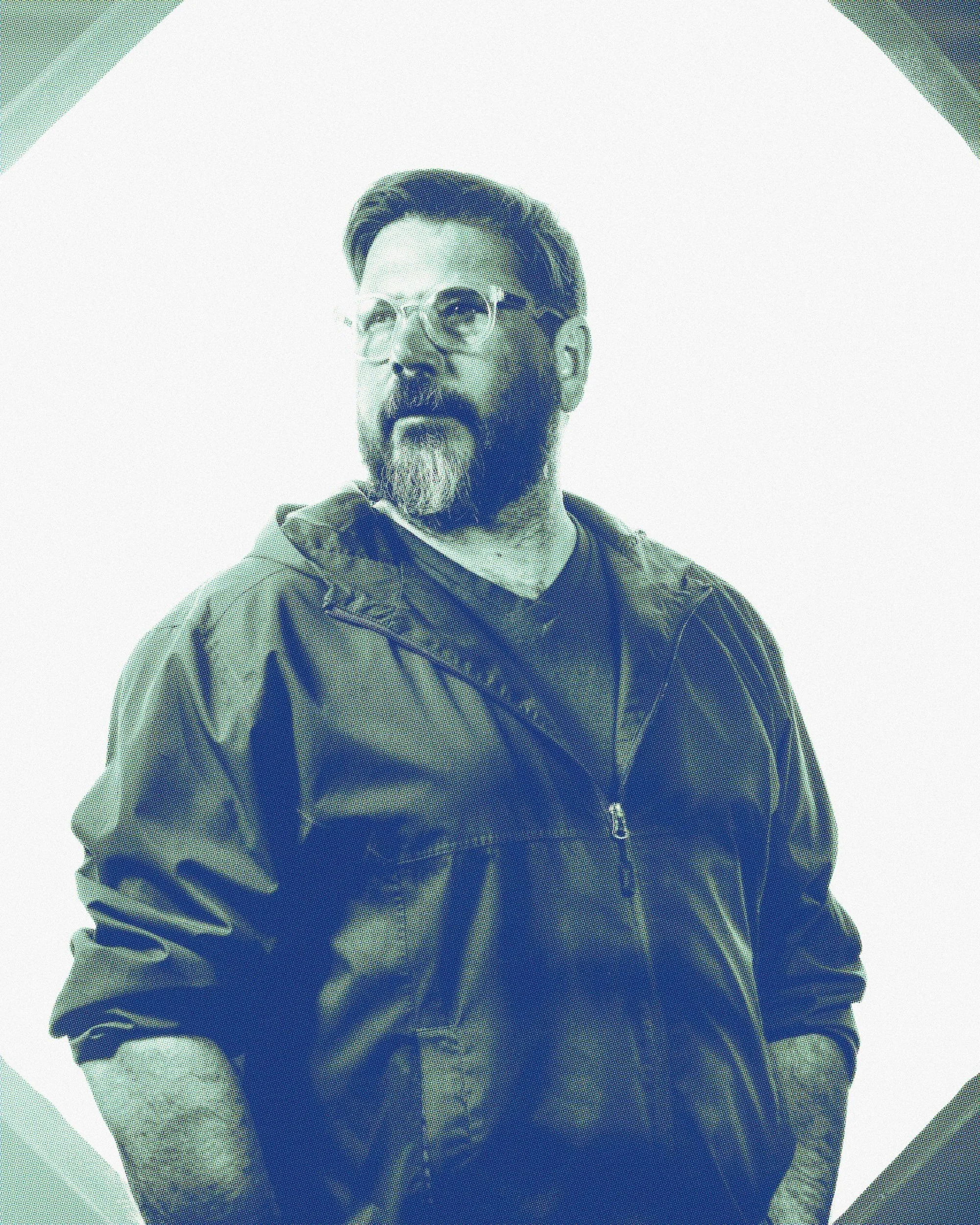Portrait of a man with glasses, a beard, and mustache, wearing a jacket, standing against a light background.
