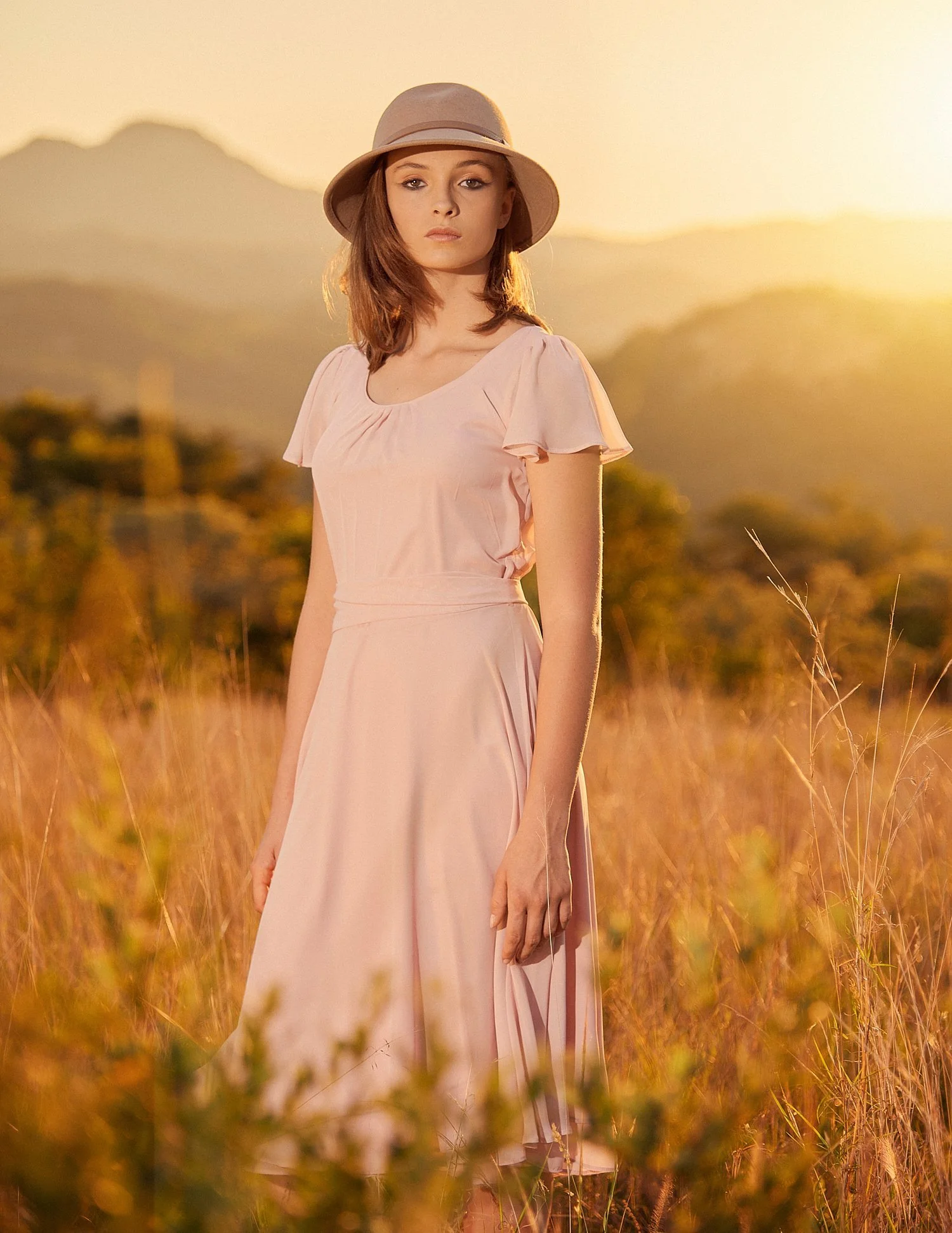 Woman in a pink dress and hat standing in a field at sunset.
