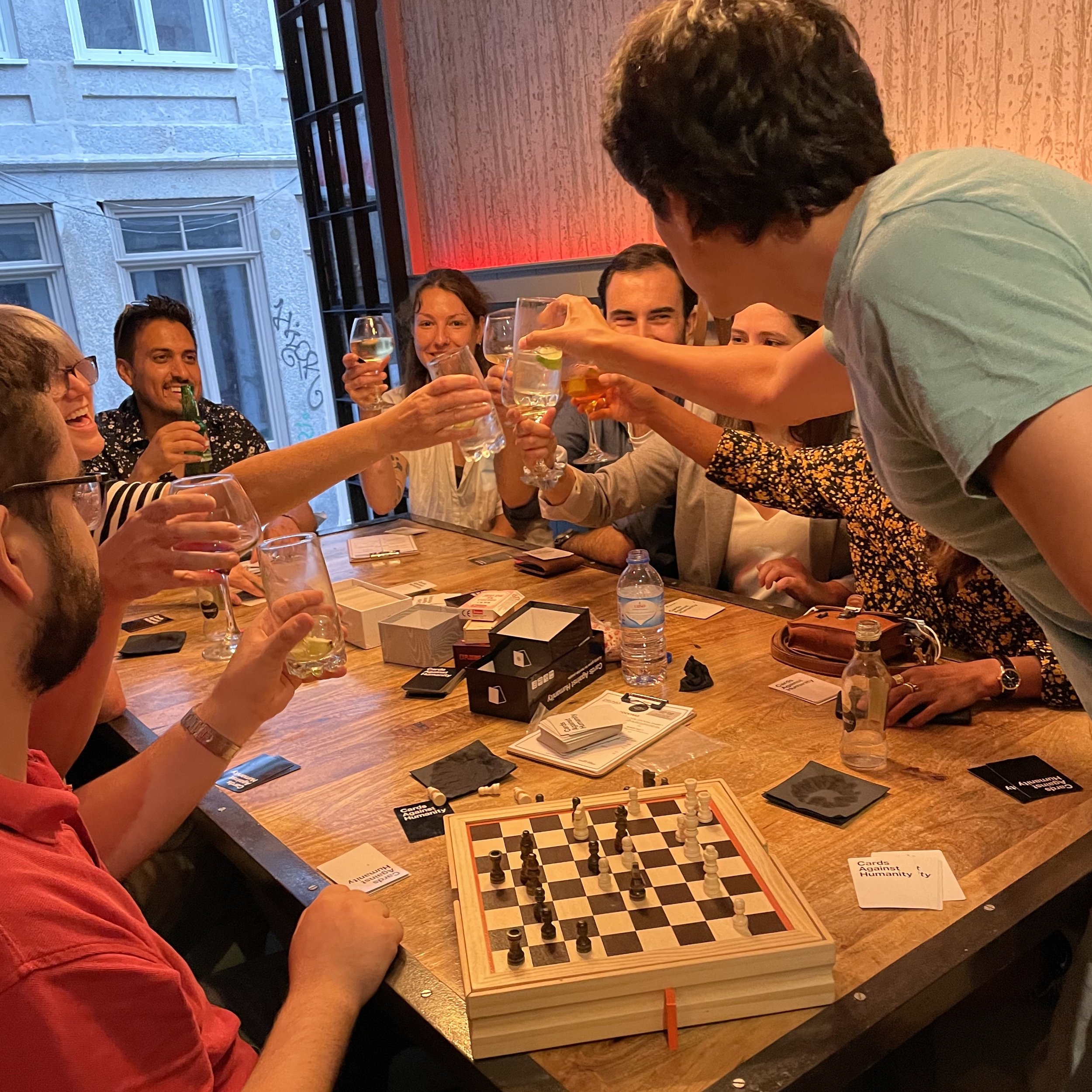 Group of people sitting around a table, holding up drinks in a toast, with a chessboard and cards on the table.