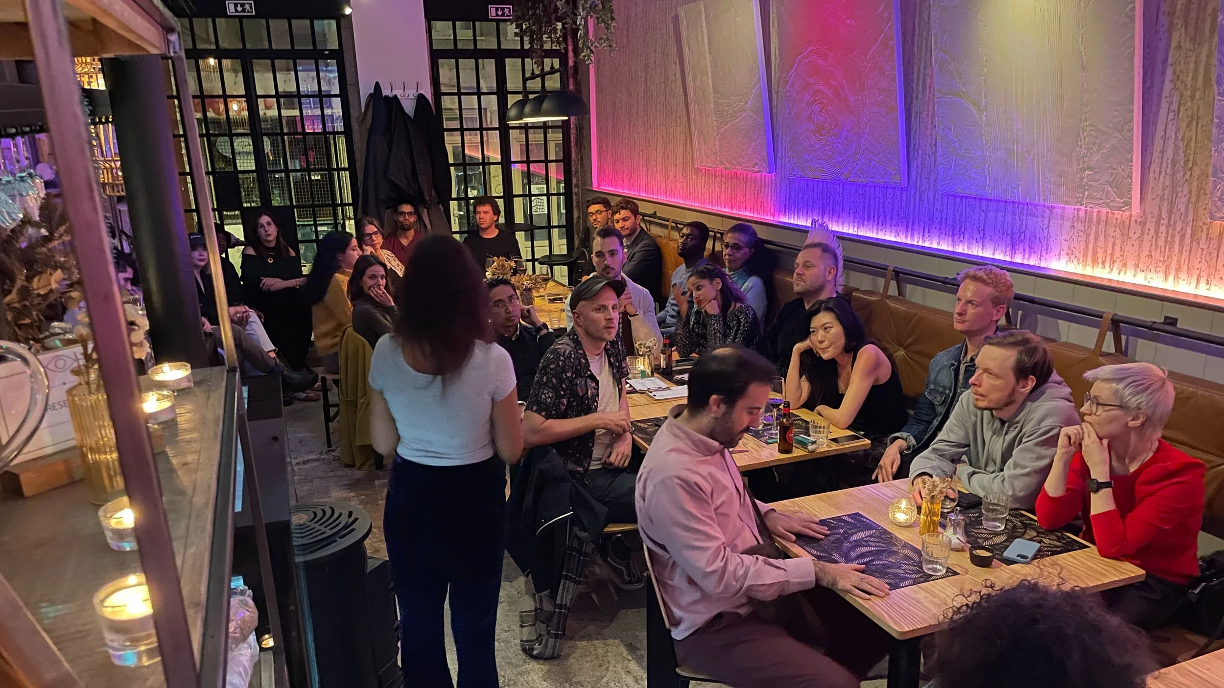 A woman speaking into a microphone at an indoor gathering, with an attentive audience seated, including a man in a floral shirt and cap. The setting has a casual, social atmosphere with dim lighting.