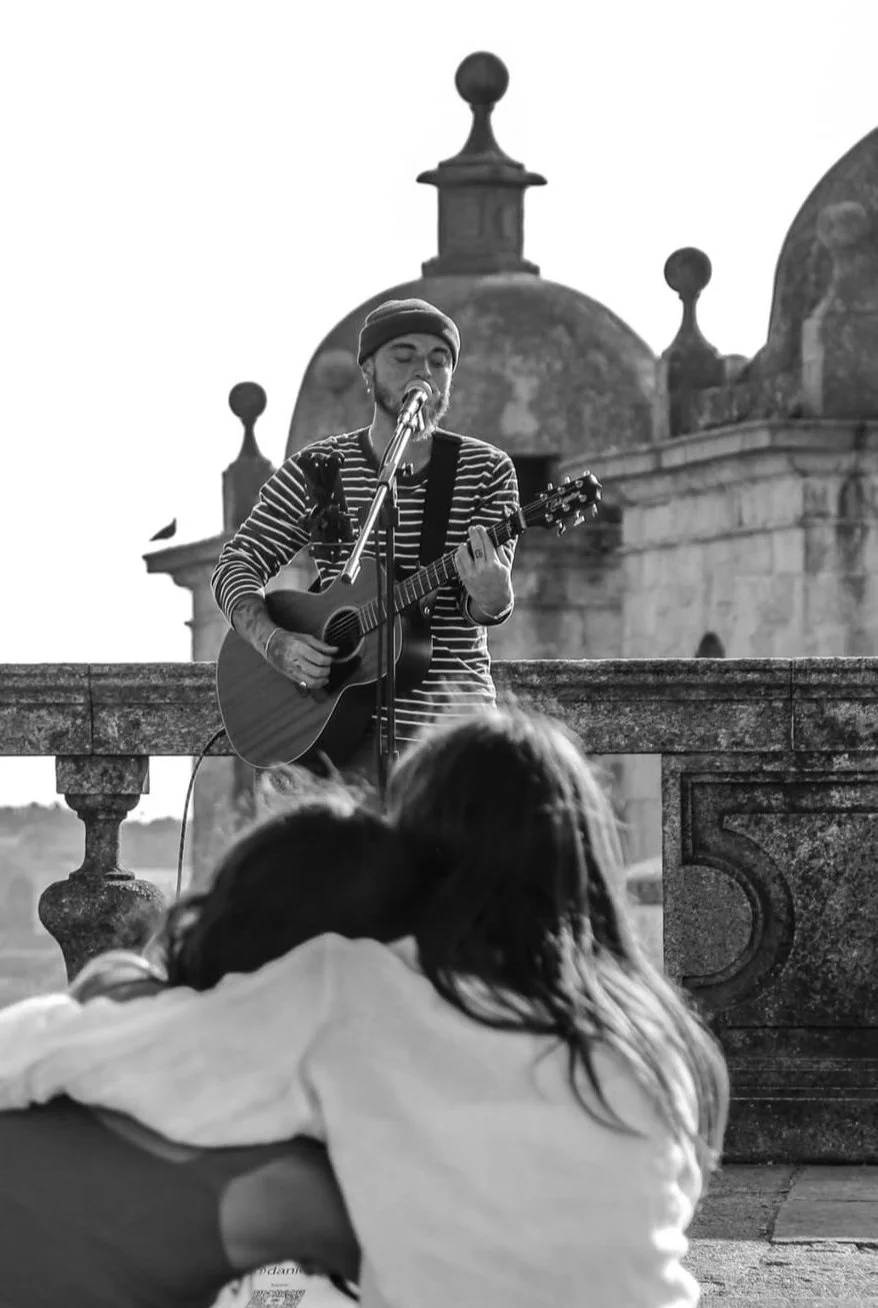 A male street musician with a beard, wearing a striped shirt and beanie, plays an acoustic guitar and sings into a microphone on a rooftop or terrace. Three people are sitting in front, watching him perform, with their backs visible. The backdrop features ornate architectural domes and balustrades, suggesting a historic or European setting, in black and white.