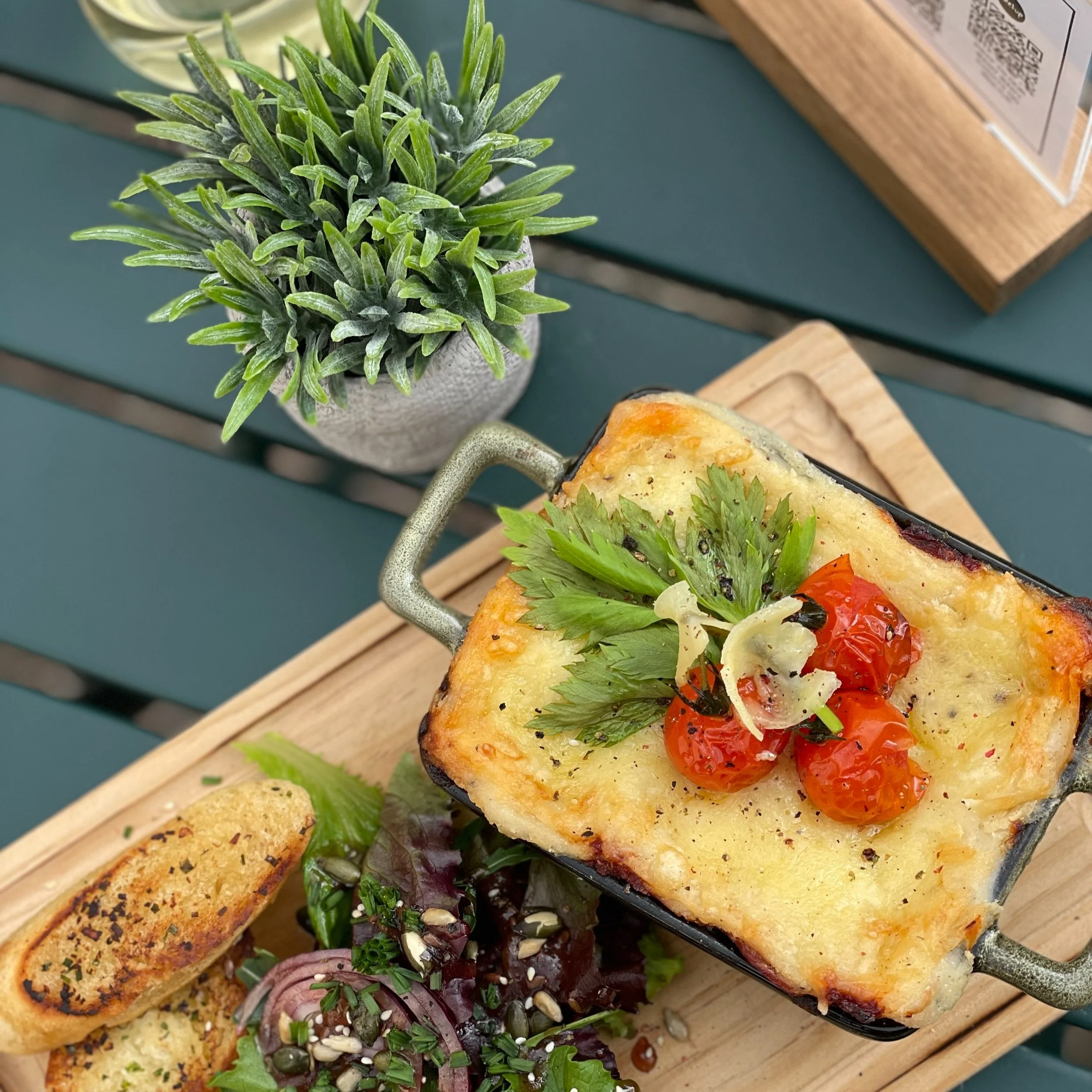 Casserole dish with melted cheese and cherry tomatoes, side salad, and toasted bread on a wooden board with a potted plant.