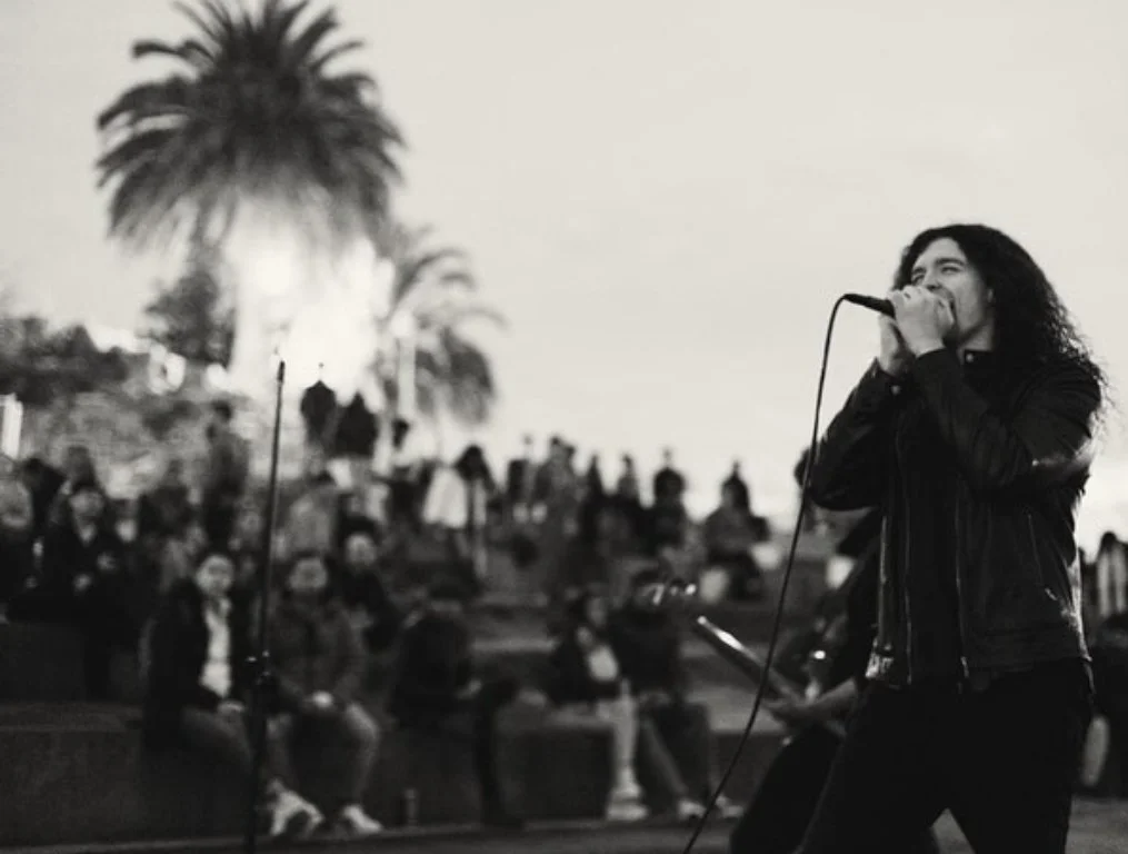 Black and white image of a musician performing outdoors with a crowd seated on steps in the background.