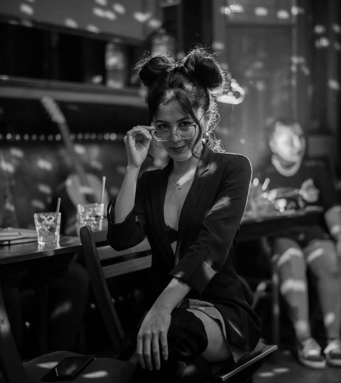 Black and white photo of a woman with glasses sitting in a cafe, wearing a blazer and knee-high boots, with ambient lighting and a guitar in the background.