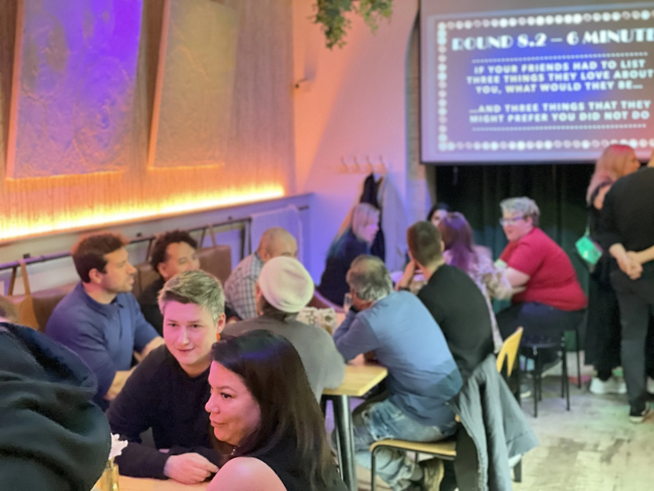 A group of people seated at tables in a cozy, dimly lit cafe or bar, engaged in conversation and participating in an activity displayed on a screen. The screen has text related to a discussion activity involving listing personal traits. The ambiance is warm with decorative elements and soft lighting.