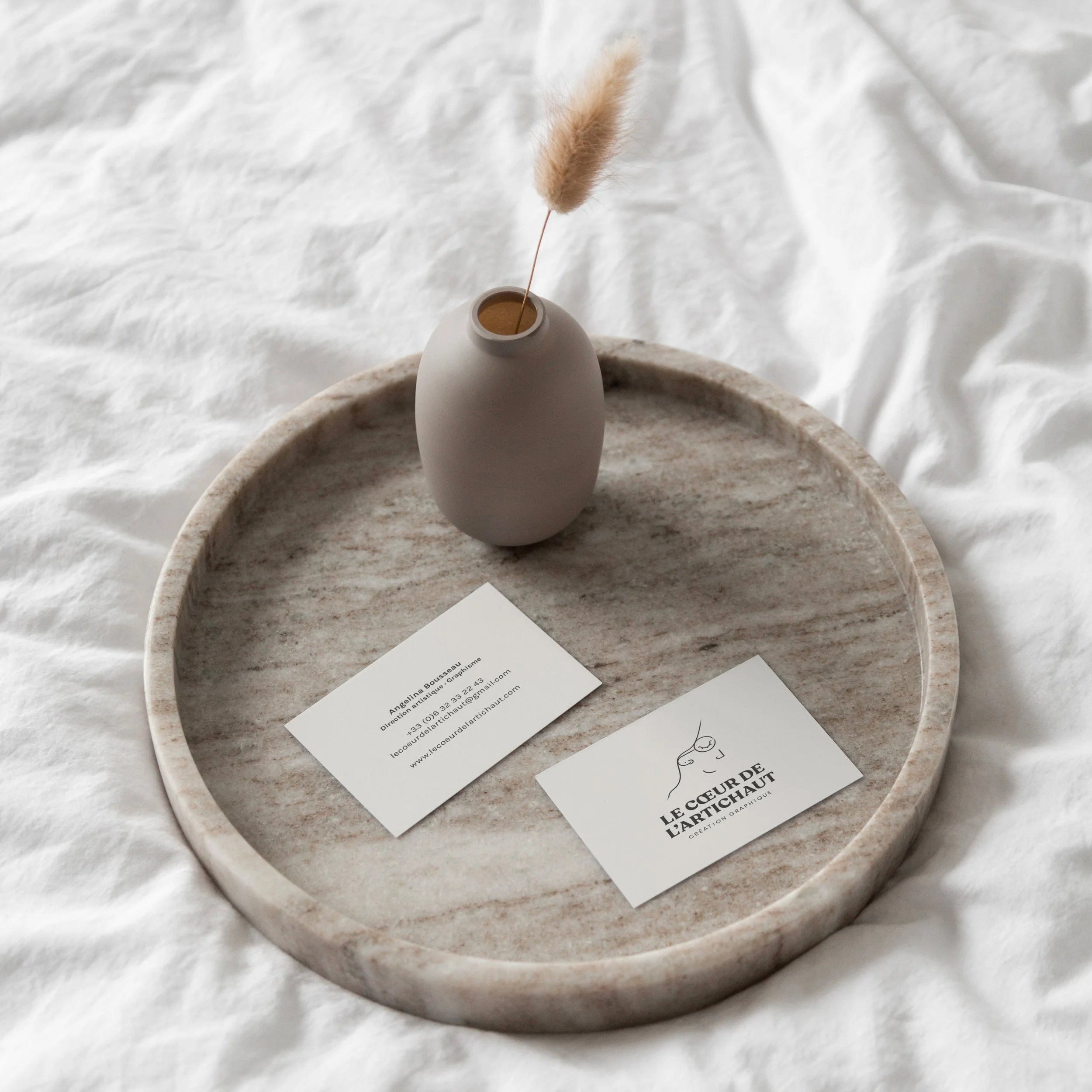Un plateau en pierre avec une petite étagère en céramique beige, une carte de visite et un autre support avec un logo et du texte, un vase beige avec une tige de pampas