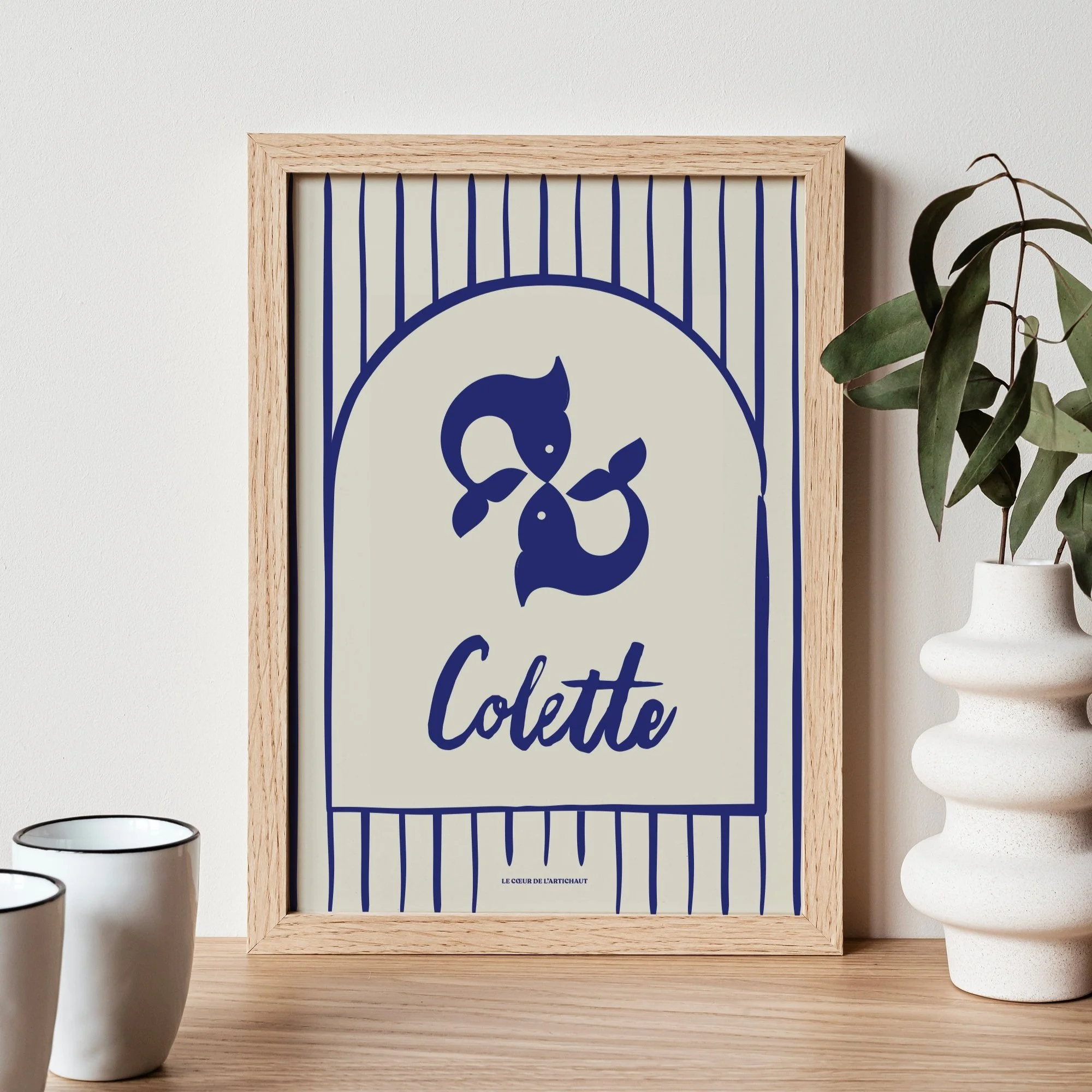Une affiche avec un dessin stylisé d'un poisson et le nom « Colette », encadrée en bois, posée sur une surface en bois avec une plante verte dans un vase blanc à droite et deux tasses blanches à gauche.