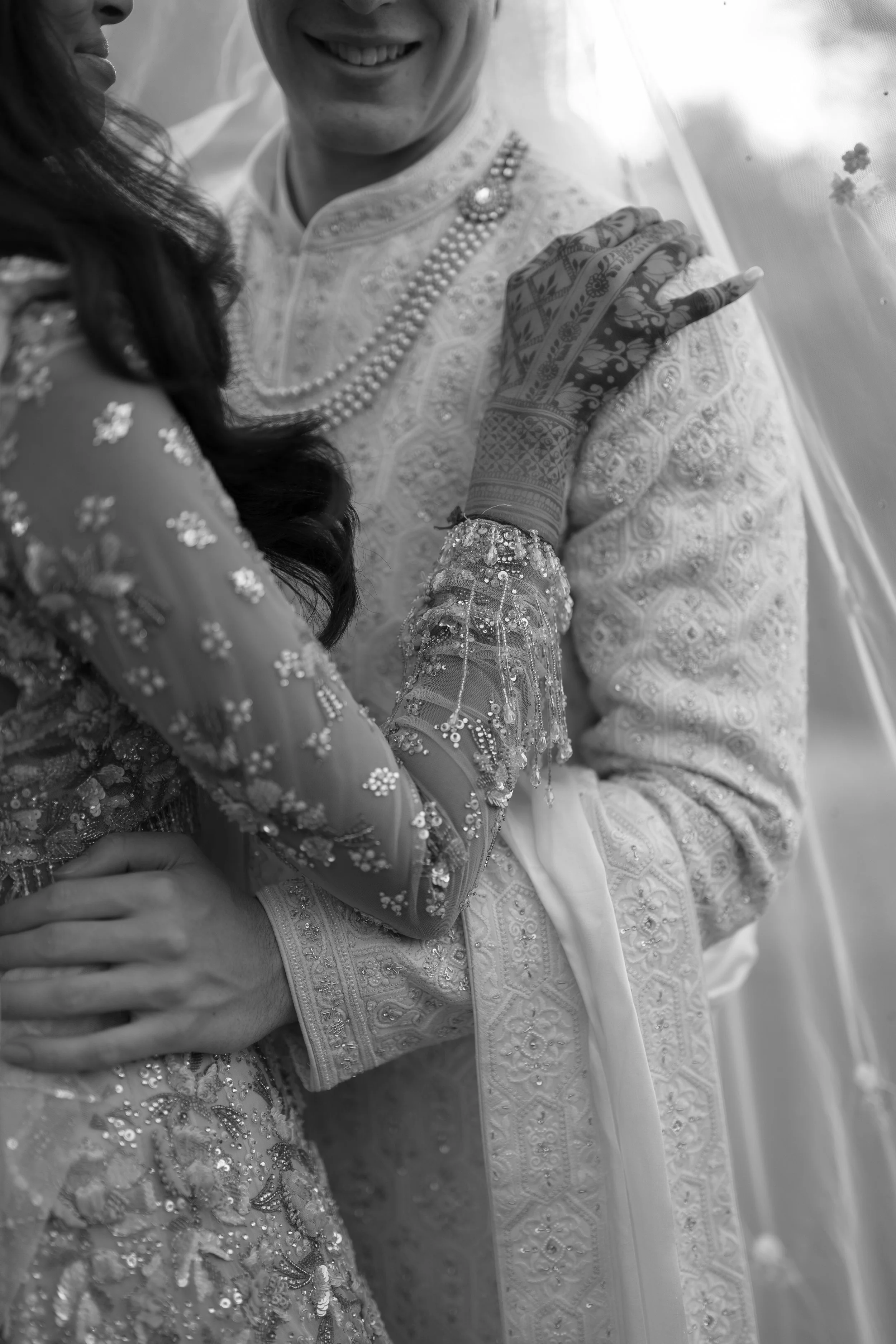 A black and white photo of an Indian bride and groom in traditional wedding attire, close together, holding hands, with detailed embroidery and jewelry.
