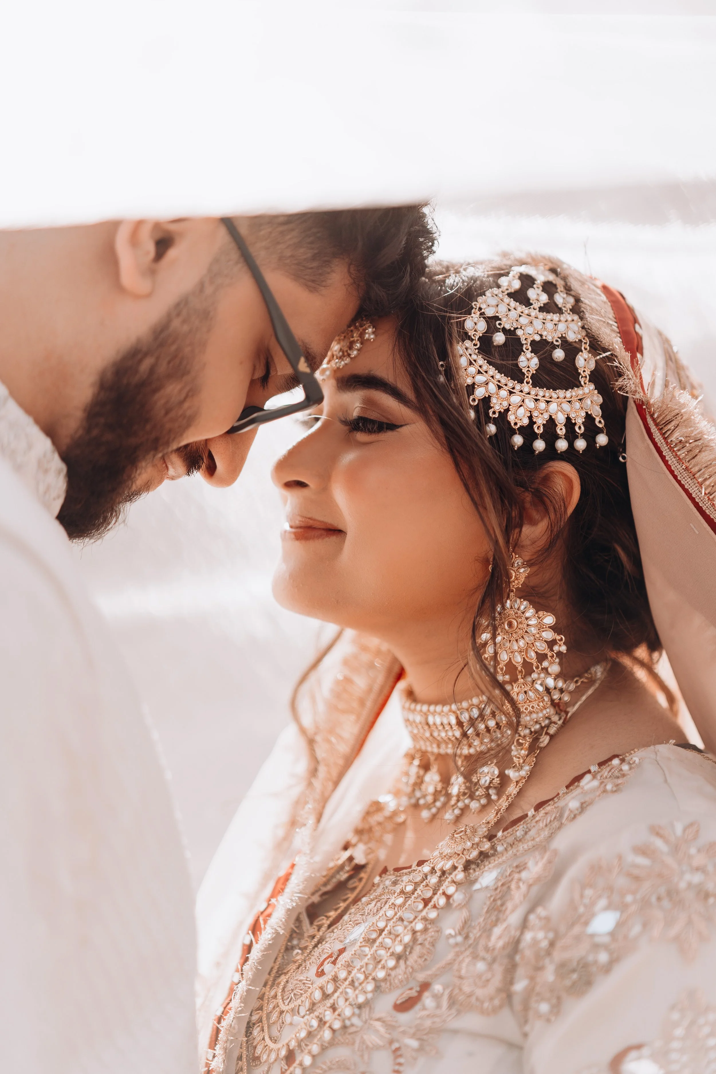 Close-up of a Pakistani bride and groom with foreheads touching, smiling, with ornate jewelry and traditional attire.