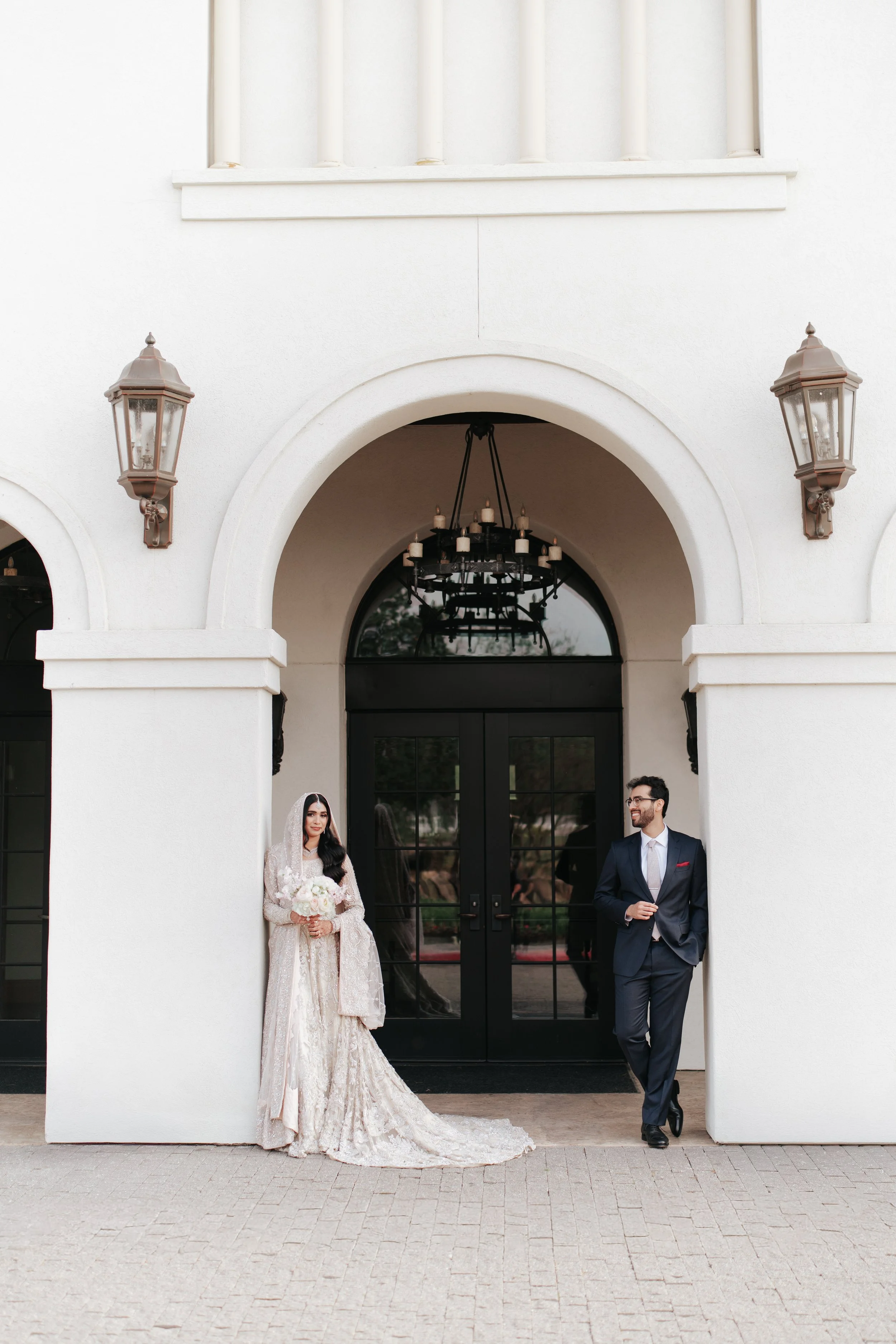 A Pakistani bride in a white wedding gown holding a bouquet and a groom dressed in a dark suit standing under an archway outside a building with white walls and lantern-style wall lights.