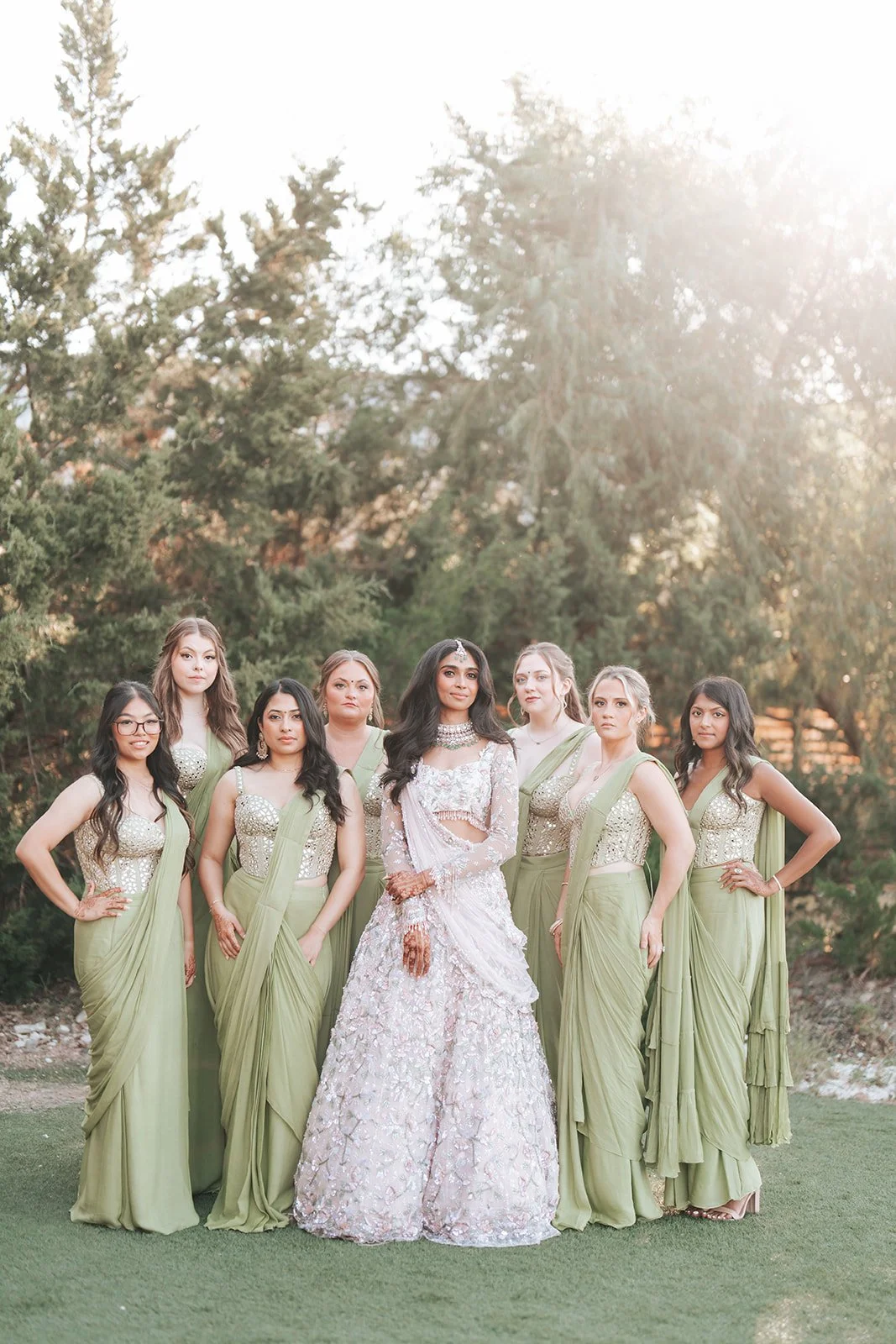 A woman in traditional Indian attire, surrounded by seven women wearing matching green and beige dresses, standing outdoors on grass with trees in the background and sunlight shining through the trees.