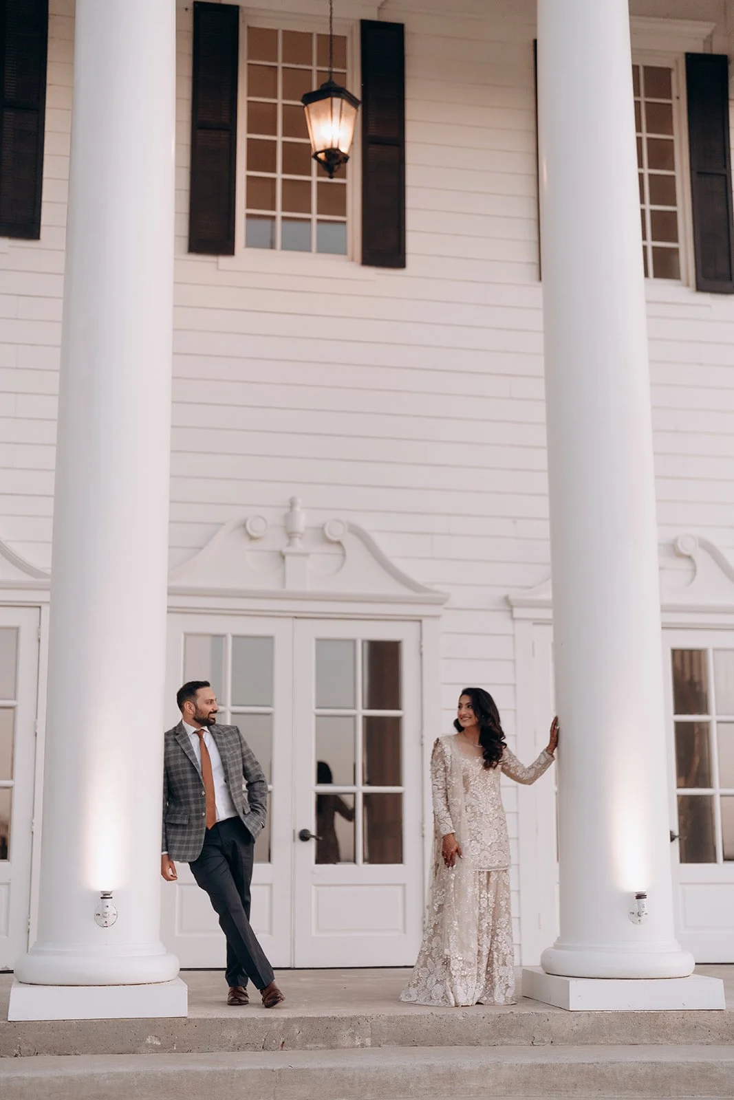 A Pakistani and Ismaili man and woman standing on the porch of a white house with large columns, engaged in conversation. The woman is wearing a light-colored, lace dress, and the man is dressed in a checkered blazer with dark trousers.