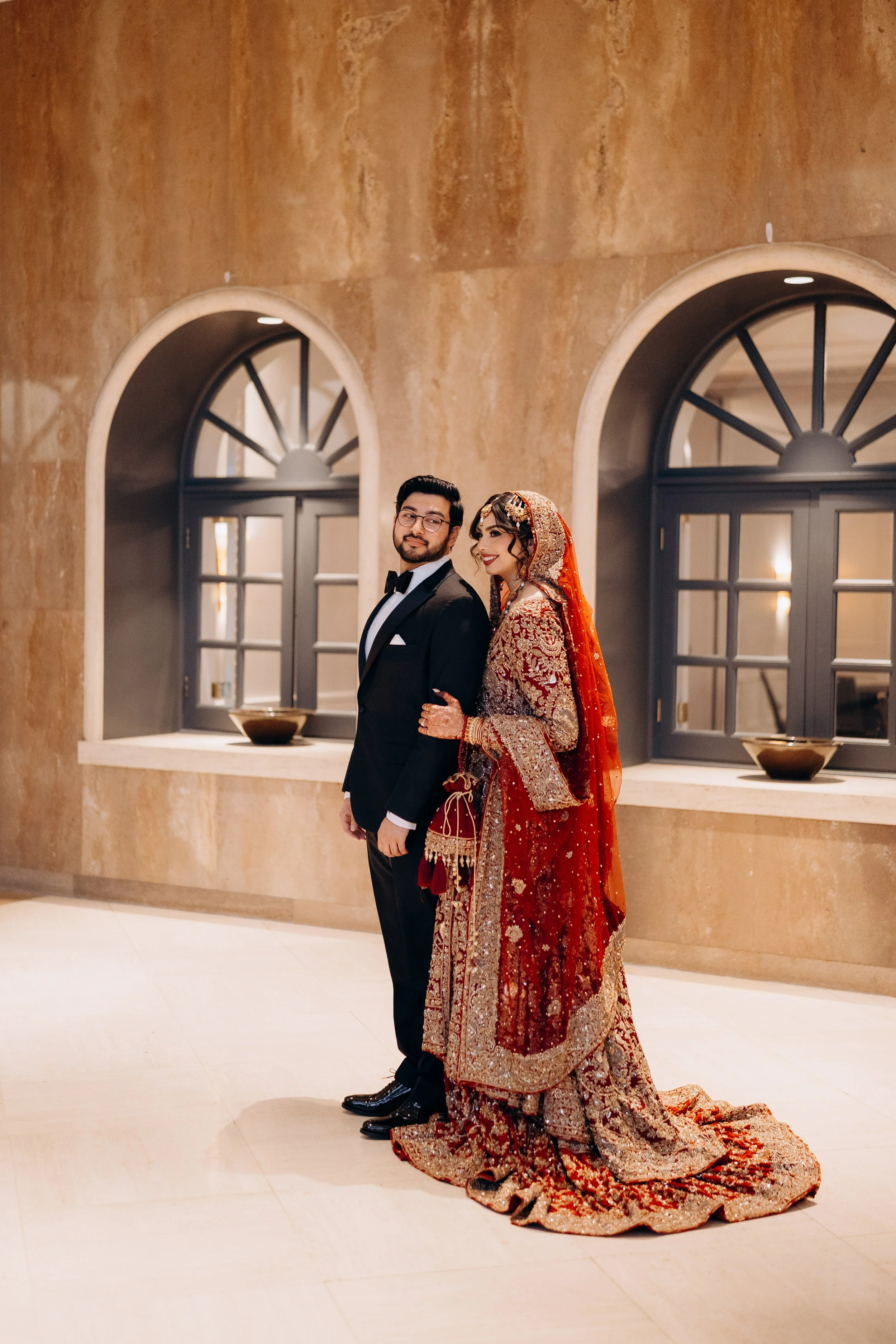 A Pakistani bride and groom standing together indoors, with the bride wearing a red and gold traditional wedding dress and the groom in a black tuxedo, in front of a beige textured wall with arched windows.