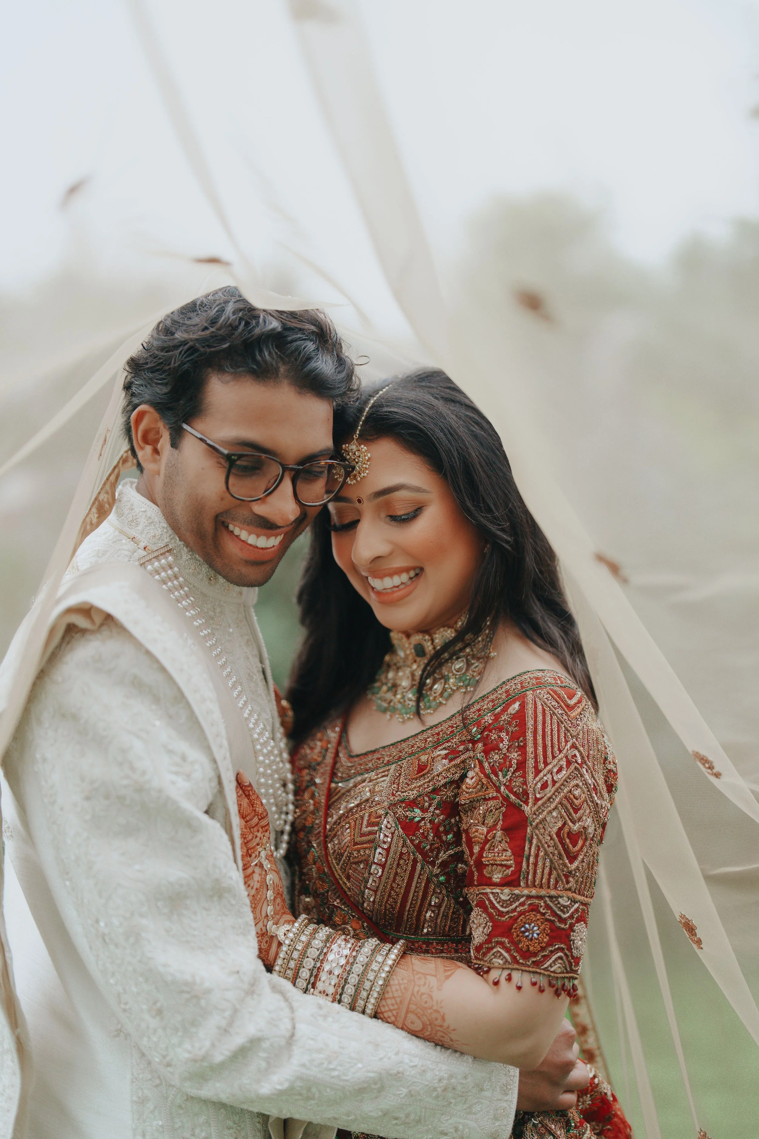 A happy Indian couple in traditional Indian wedding attire under sheer fabric, smiling and embracing.