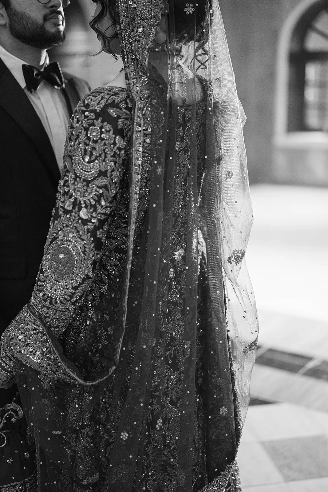 A couple in Pakistani wedding attire, the bride wearing a heavily embroidered dress with a veil covering her face, standing next to the groom in a tuxedo with a bow tie, in an indoor setting.