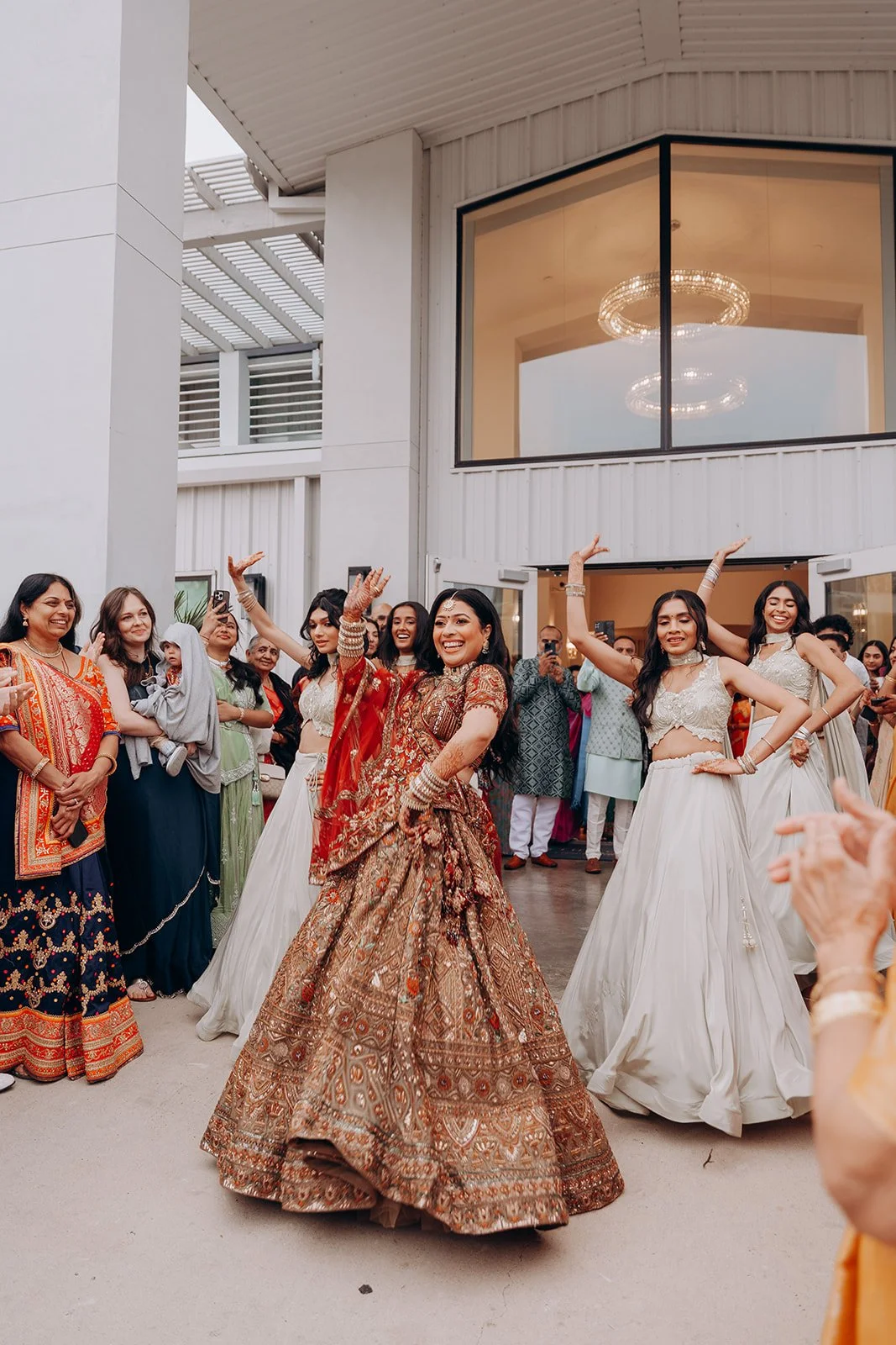Group of Indian women celebrating at a wedding, with the bride in traditional Indian attire dancing and smiling, surrounded by family and friends in colorful clothing.