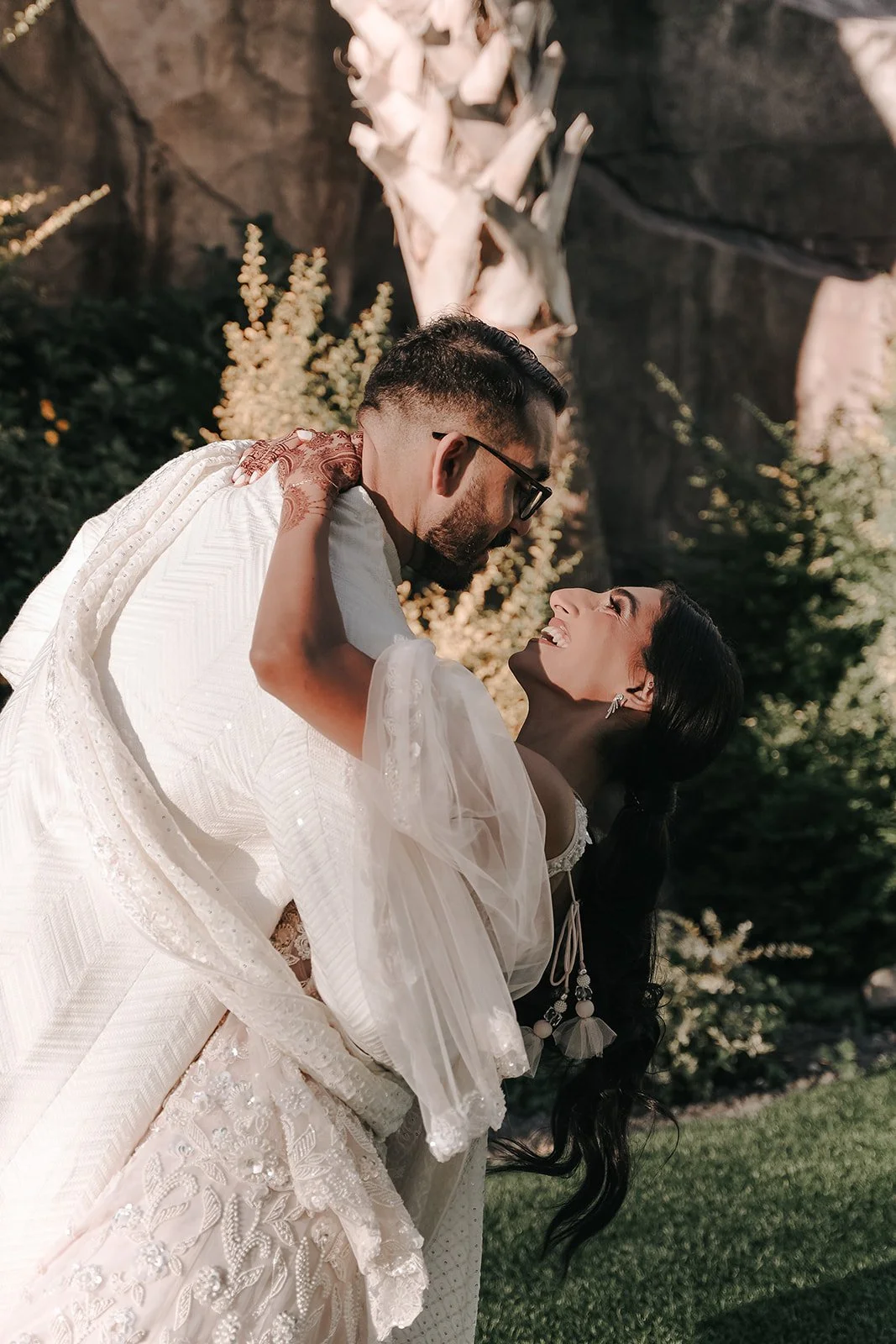 An Ismaili couple dressed in wedding attire, with the man holding the woman in a dip, smiling back at each other outdoors with mountains and greenery in the background.