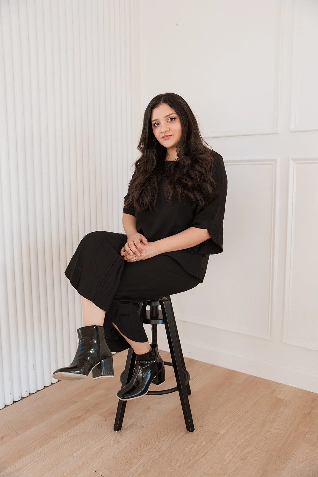 A Pakistani woman with long hair sitting on a black stool against a white wall, wearing a black outfit and black shiny ankle boots.