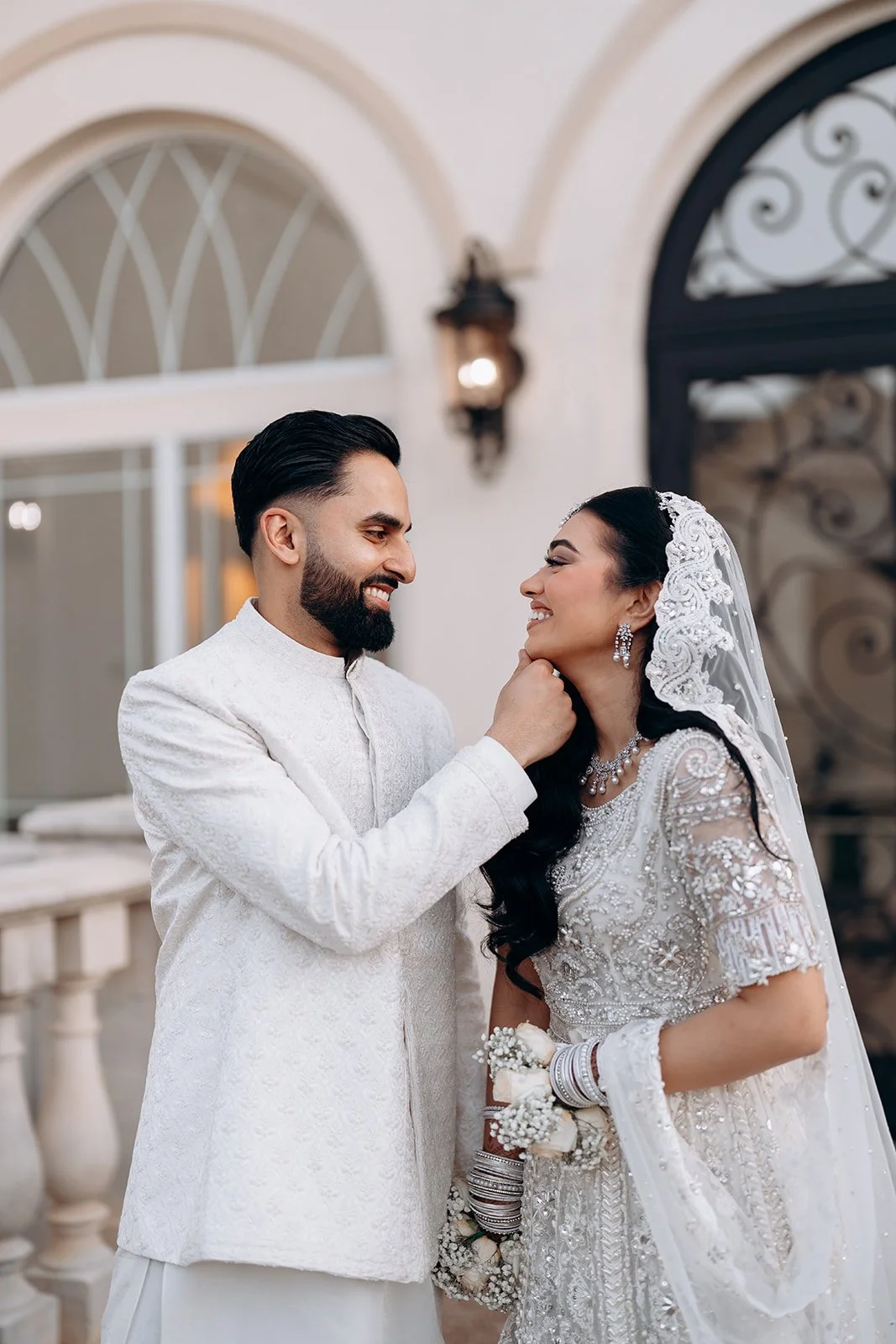 A Pakistani groom and bride smiling at each other, with the groom holding the bride's chin, outside a building with arches and ornate lattice windows.