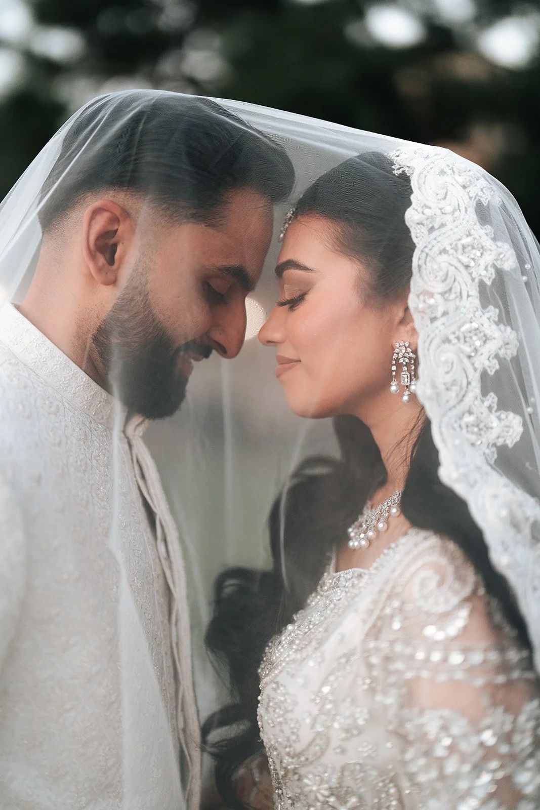 A Pakistani bride and groom with foreheads touching, smiling softly under a wedding veil, dressed in traditional wedding attire.