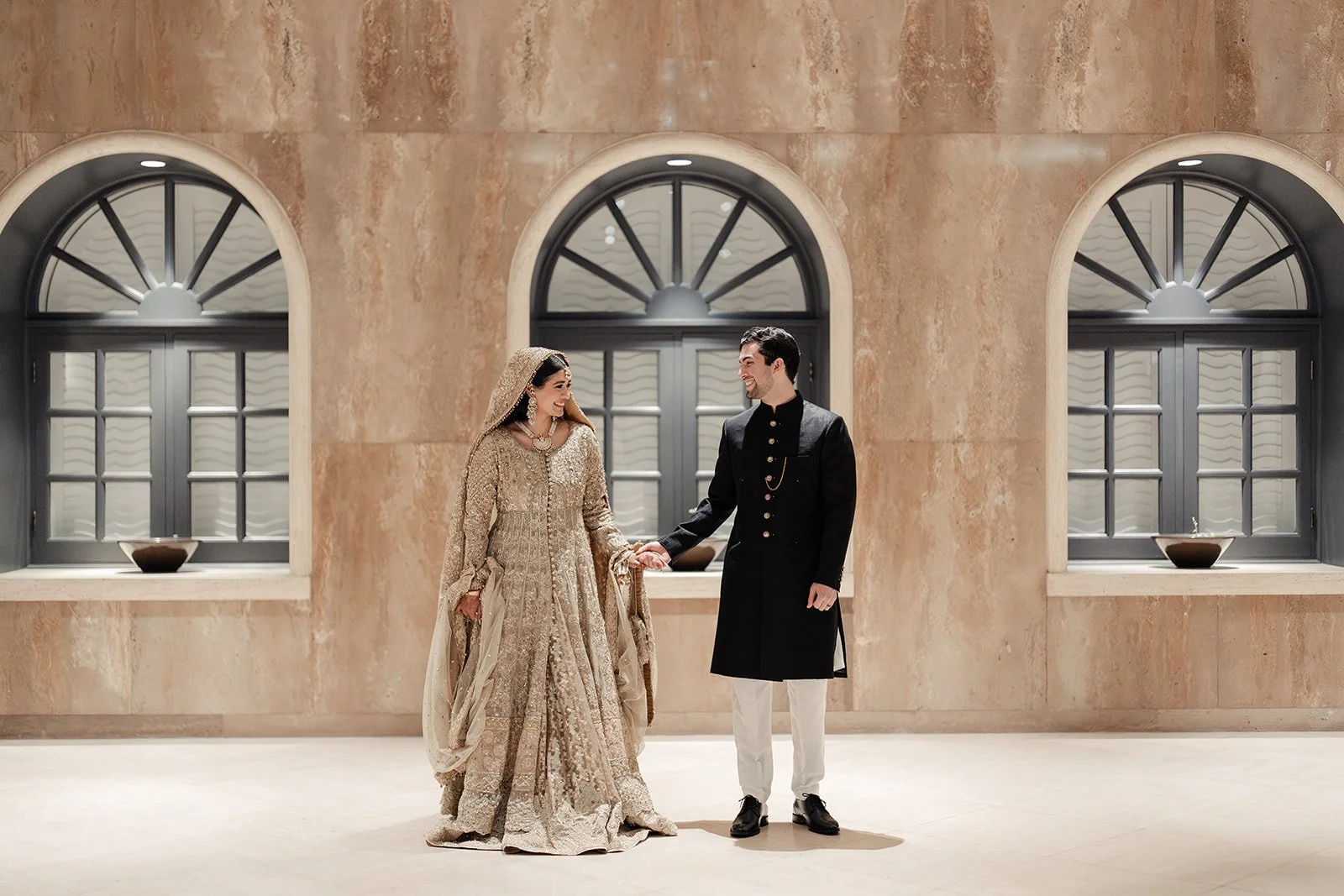 A Pakistani bride and groom dressed in traditional Indian wedding attire holding hands and smiling at each other inside a modern hall with three arches and large windows.
