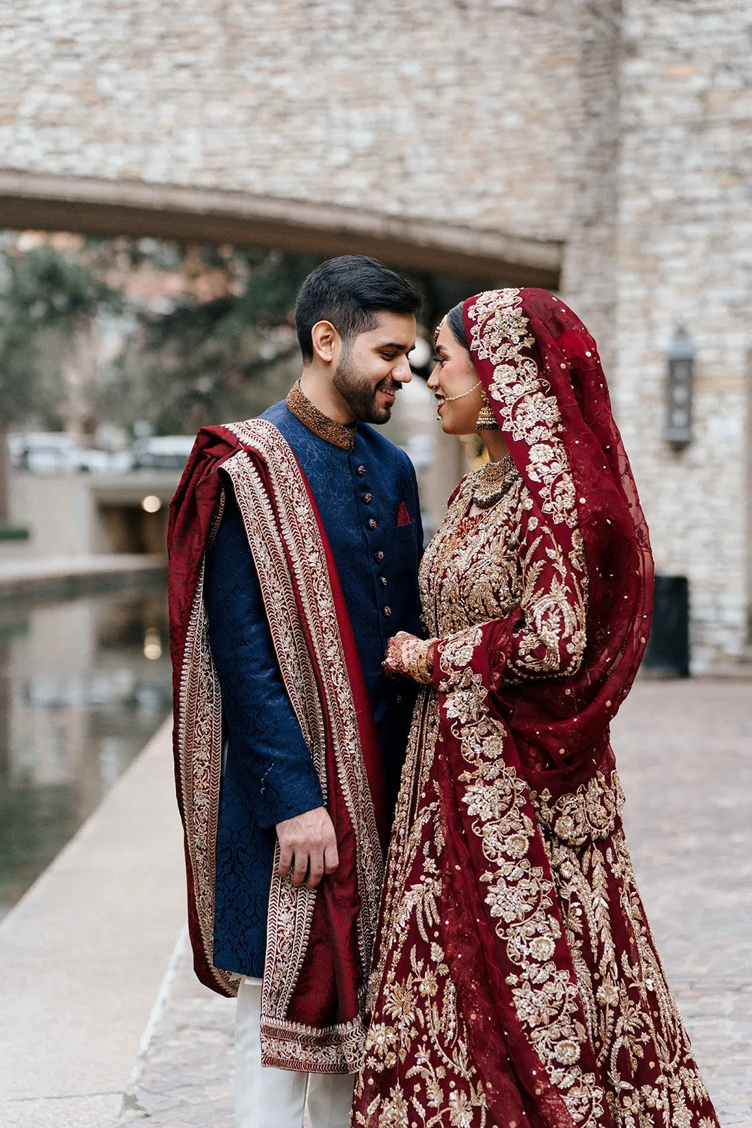 A happy Pakistani couple in traditional Indian wedding attire standing close to each other near a body of water, with a stone building and trees in the background.